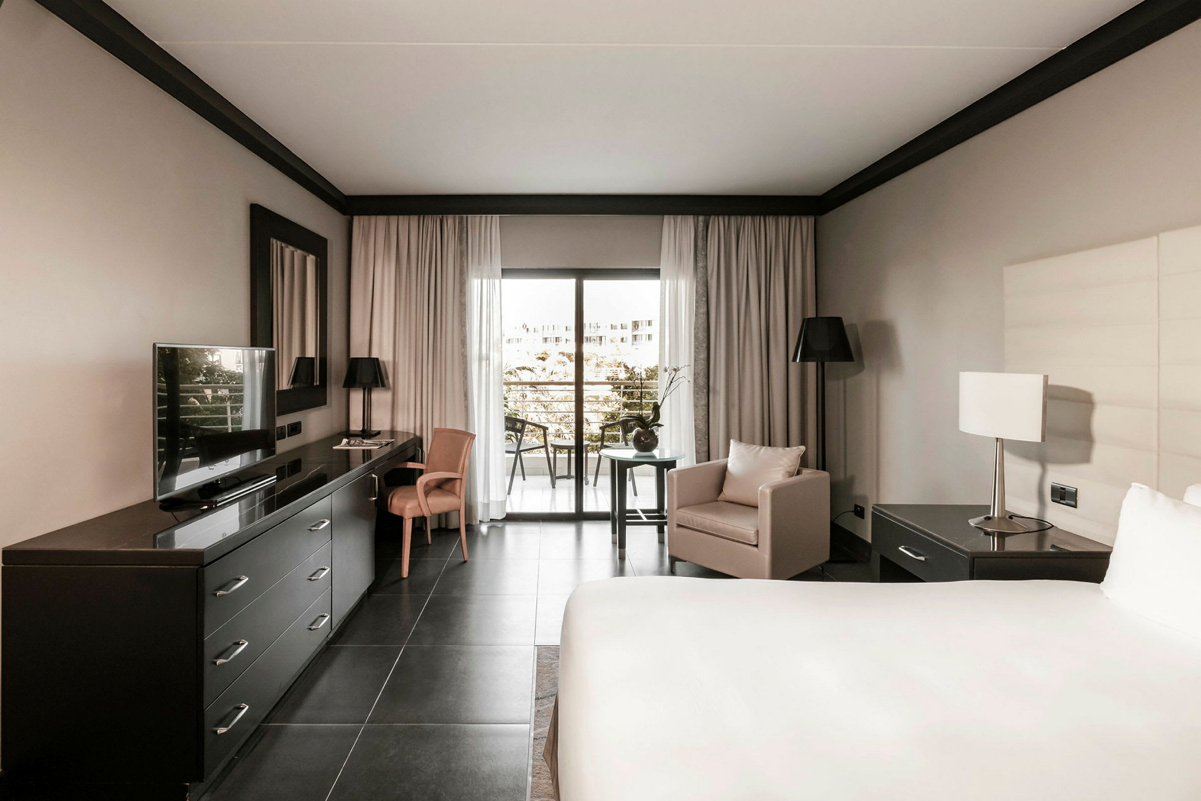 Hotelværelse med moderne indretning, en stor seng, skrivebord med TV, lænestol og vindue mod en balkon. (AI-genereret tekst)