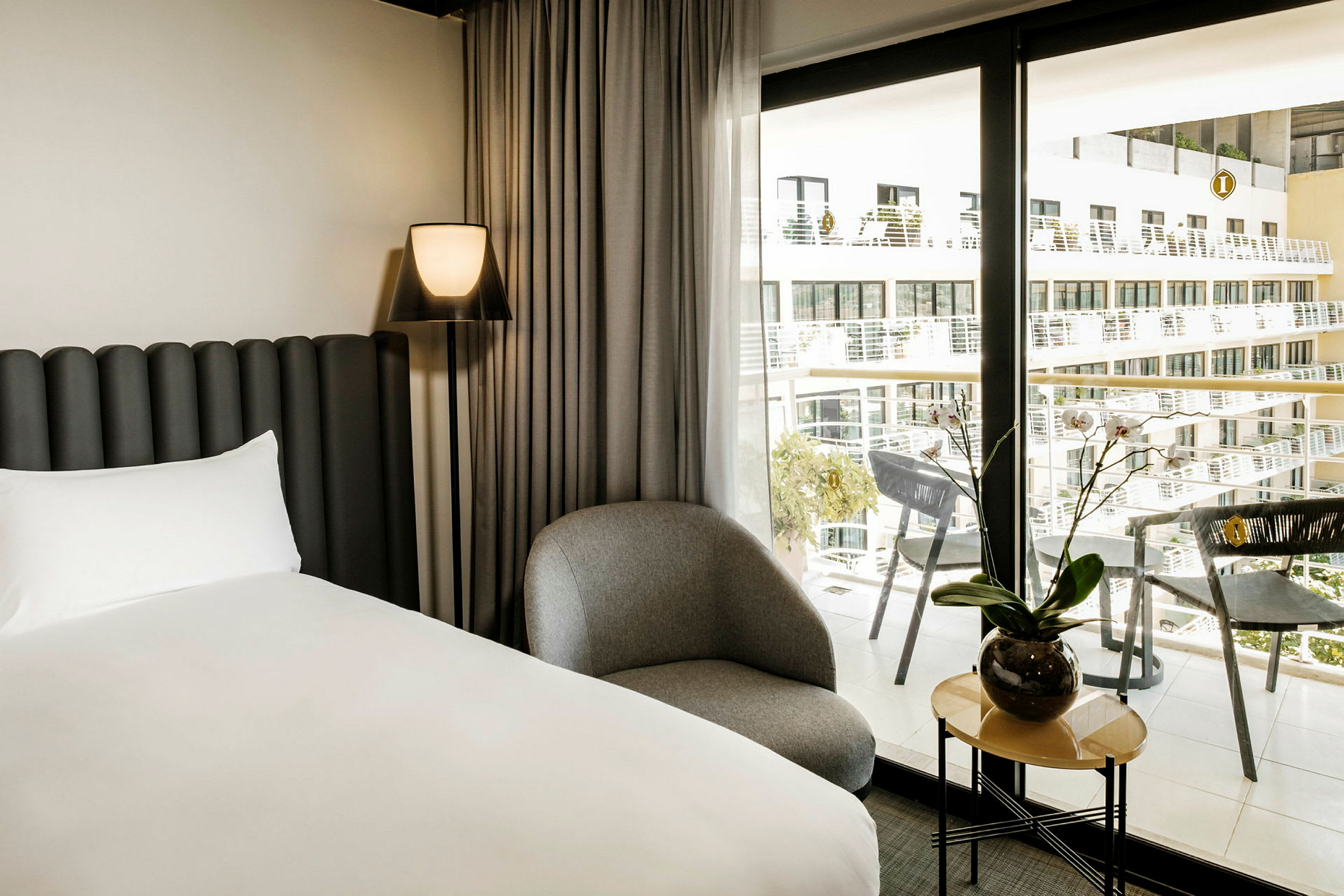 Et moderne hotelværelse med en seng, en stol og et lille bord med en plante ved siden af et stort vindue med udsigt over altaner. (AI-genereret tekst)