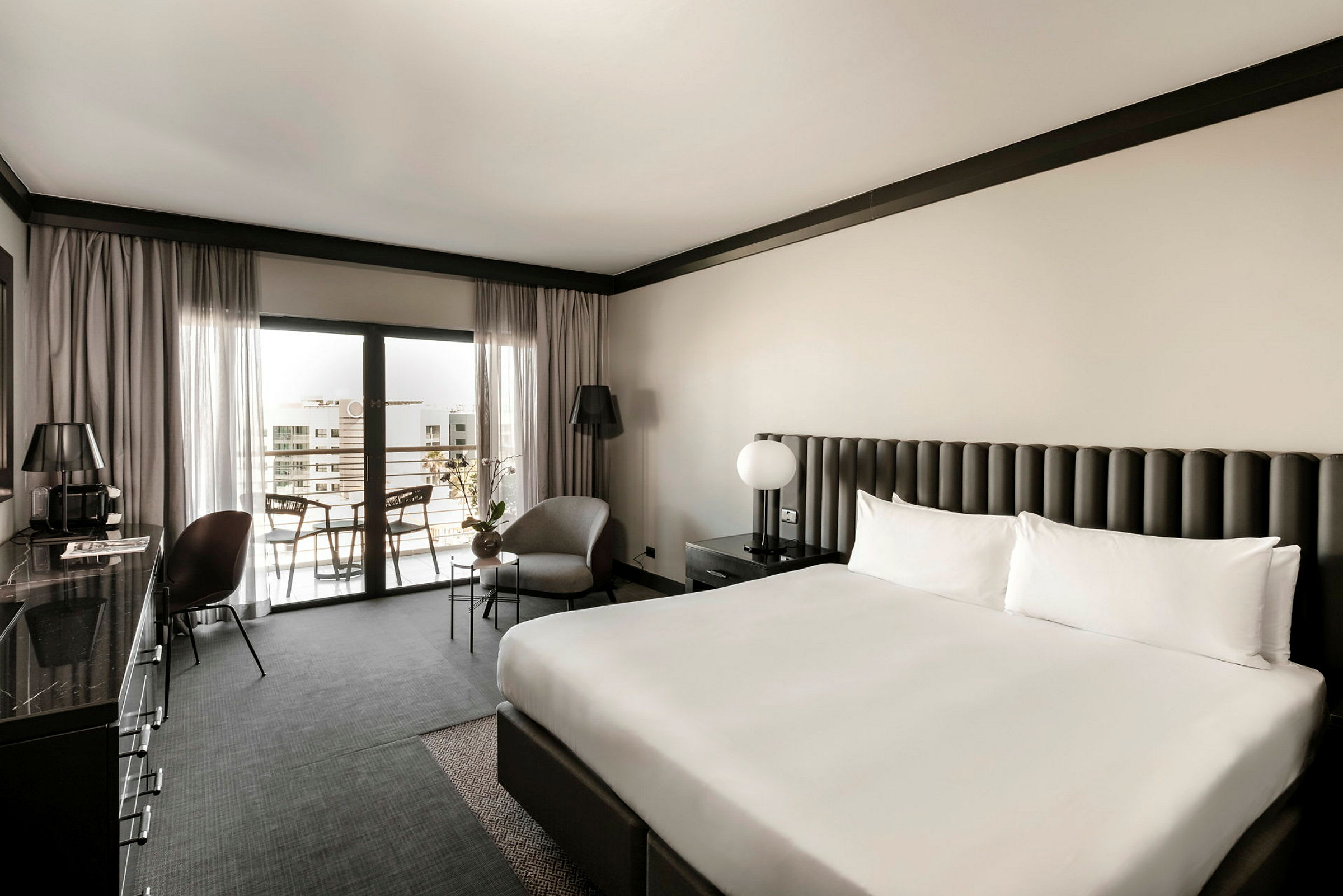 Hotelværelse med en stor dobbeltseng, lænestol og bord ved siden, samt balkonsiddeplads. Elegant neutraltonede møbler og indretning. (AI-genereret tekst)