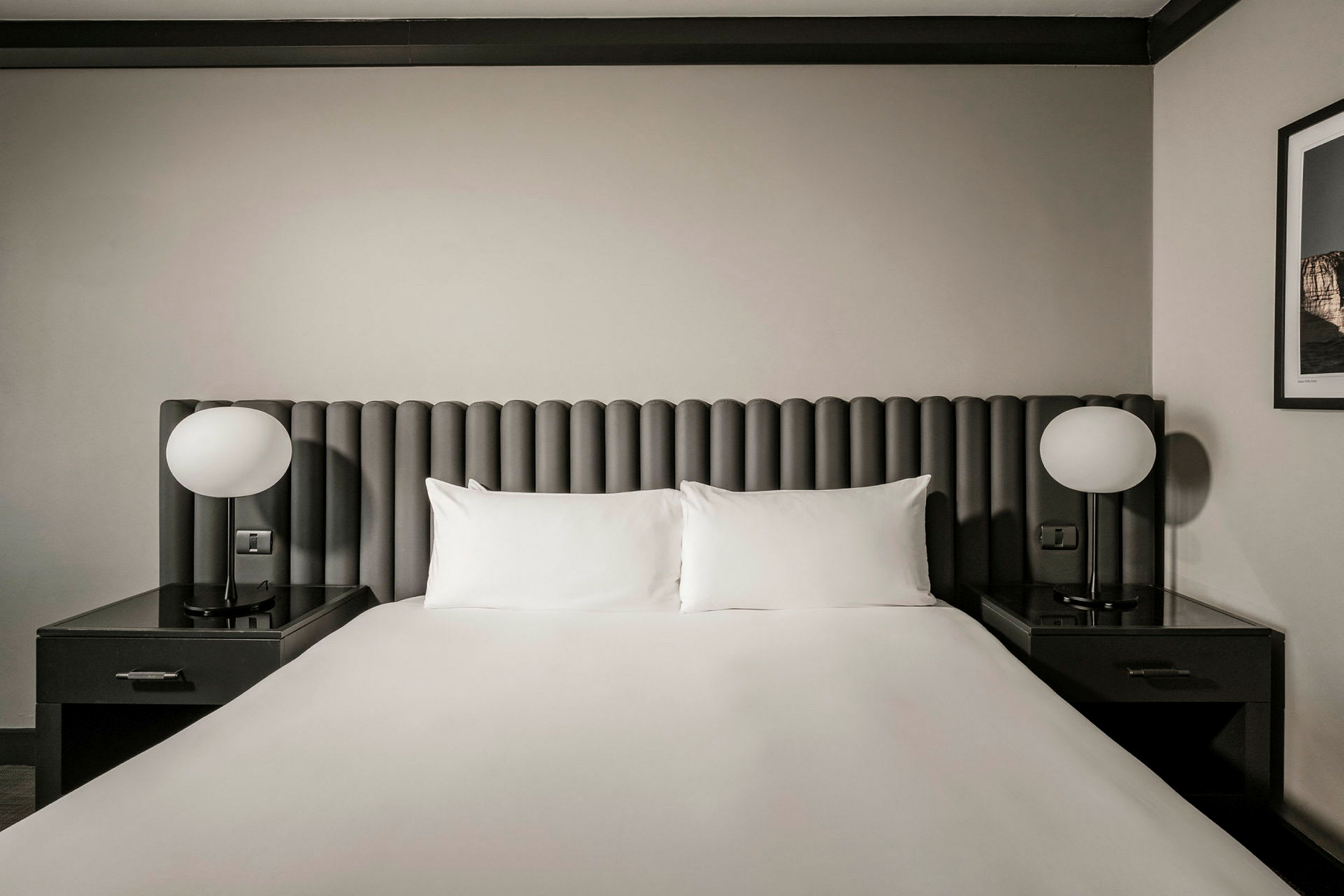 Et hotelværelse med en dobbeltseng. Sengen har en grå hovedgærde, hvide puder og lagner. Ved hver side er der natbord med runde lamper. (AI-genereret tekst)