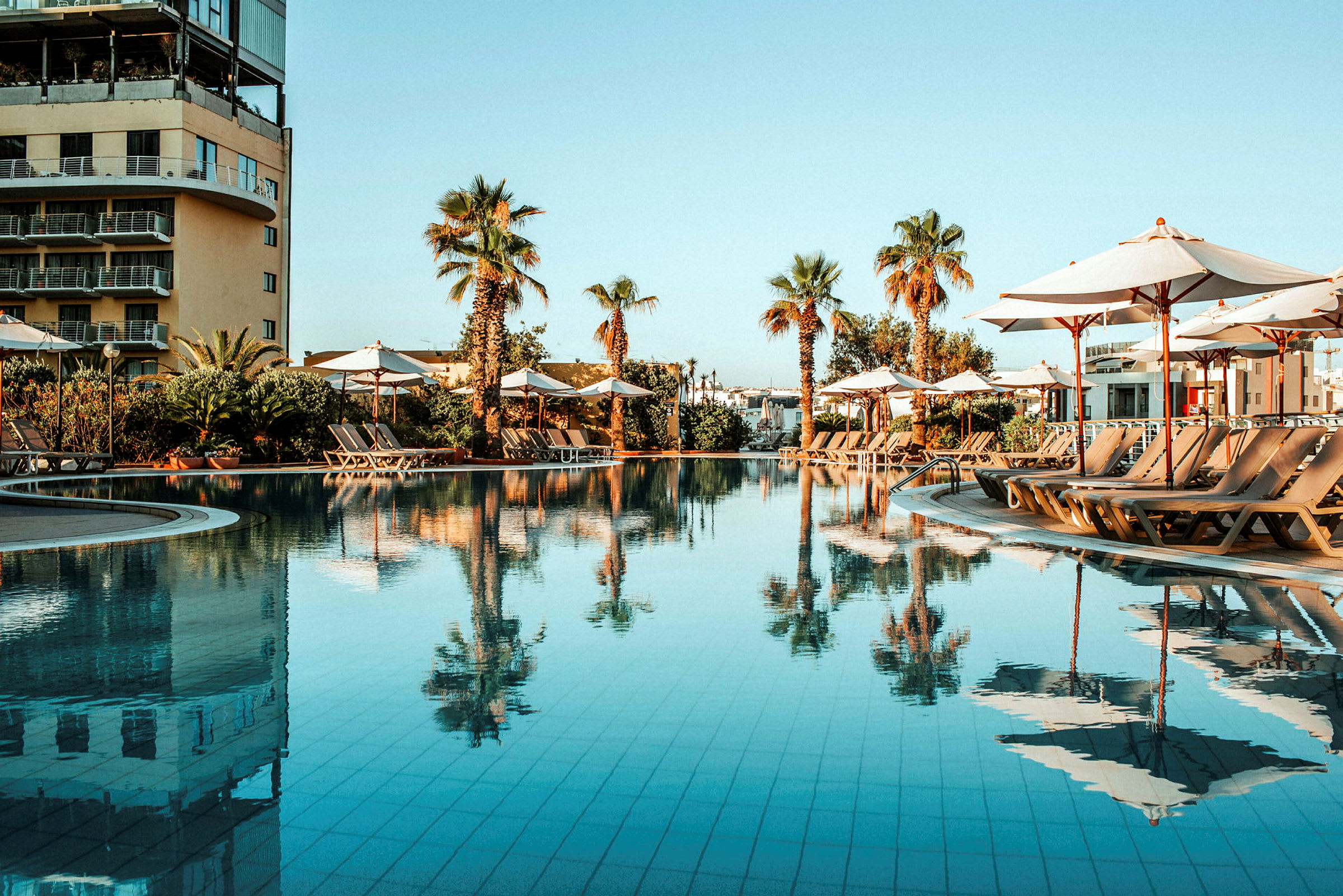 En swimmingpool med liggestole og parasoller omgivet af palmer. (AI-genereret tekst)