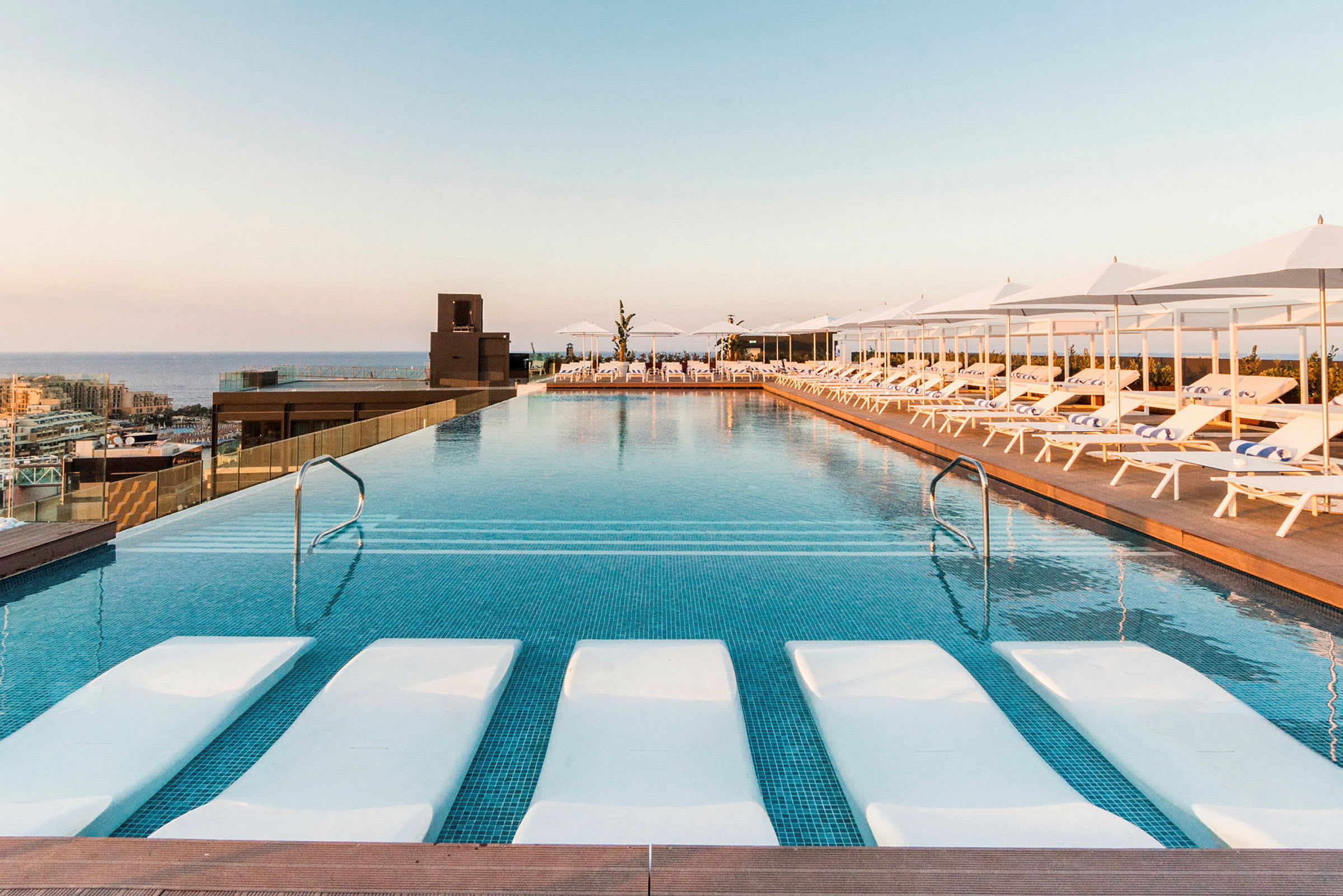 En infinity-pool med liggestole og parasoller ved siden af. Solnedgang ses i baggrunden. (AI-genereret tekst)