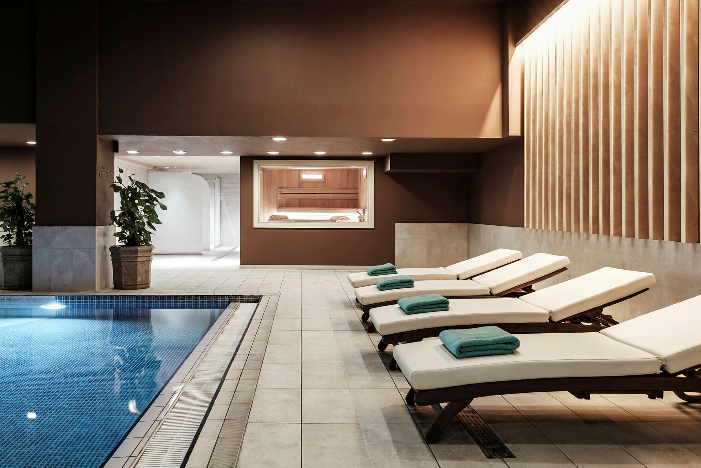 Interiørbillede af et spaområde med en pool, liggestole med håndklæder og en sauna synlig gennem vinduet. (AI-genereret tekst)