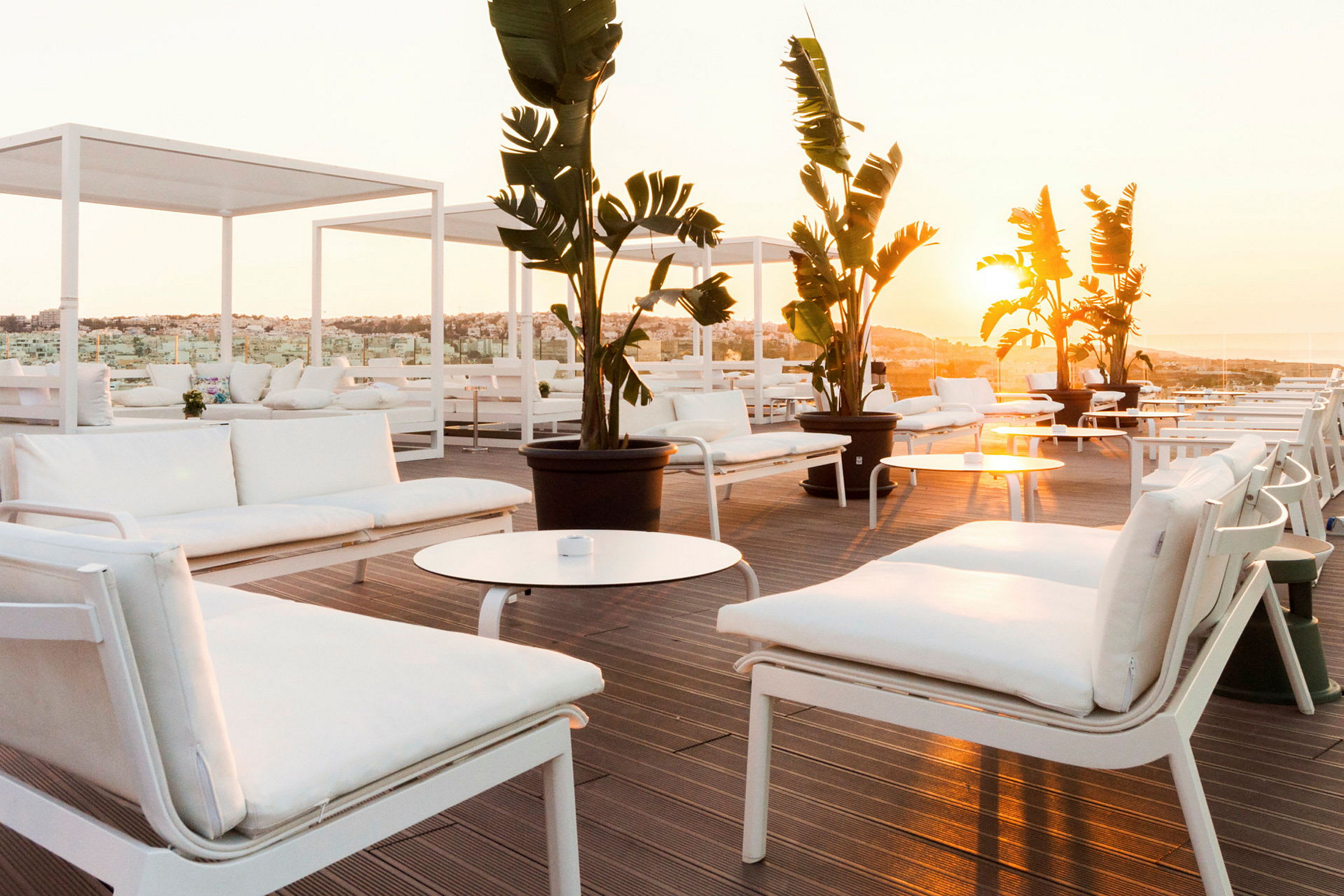 Moderne hvide sofaer og borde på en solrig terrasse med udsigt over det omkringliggende landskab, dekoreret med store potteplanter. (AI-genereret tekst)