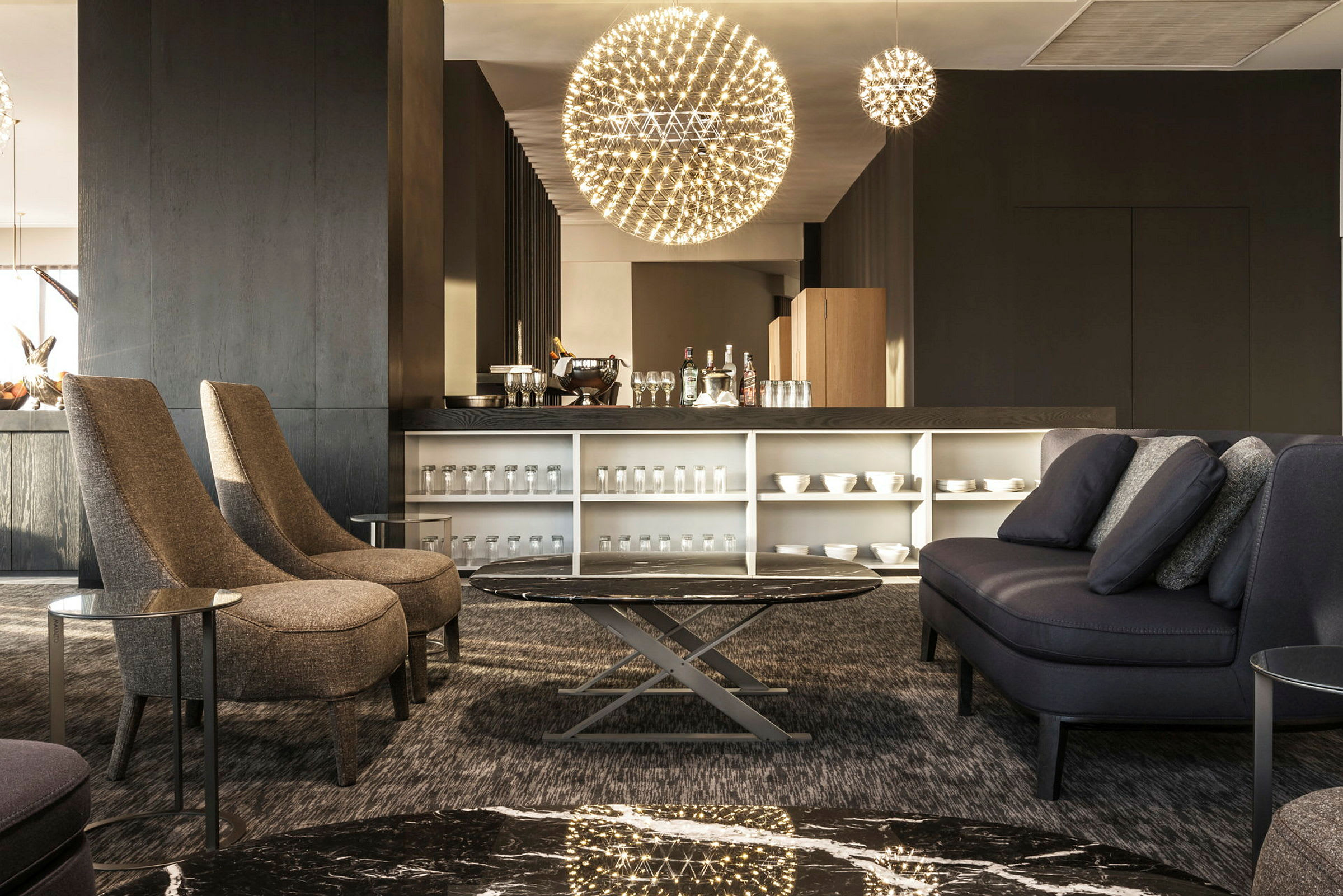 En moderne lounge med kaffebar og siddeområde med sofaer og stole. Rummet er godt oplyst med unikke hængende lamper og har et elegant indrettet bord. (AI-genereret tekst)