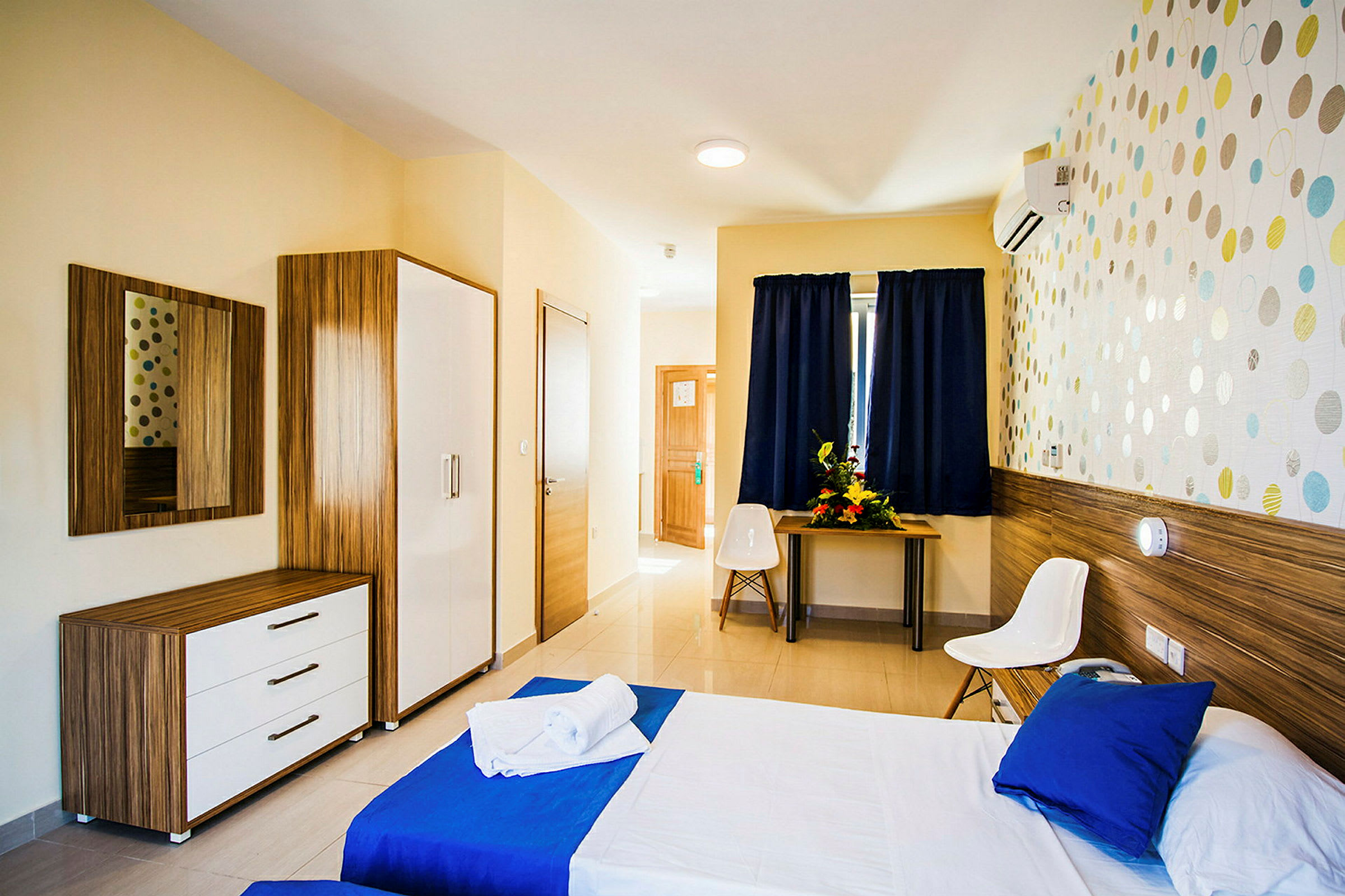 Et lyst og moderne hotelværelse med en seng redt op med blå og hvide lagner. Værelset har et spejl og møbler i træ samt et skrivebord med en blomsterdekoration. (AI-genereret tekst)