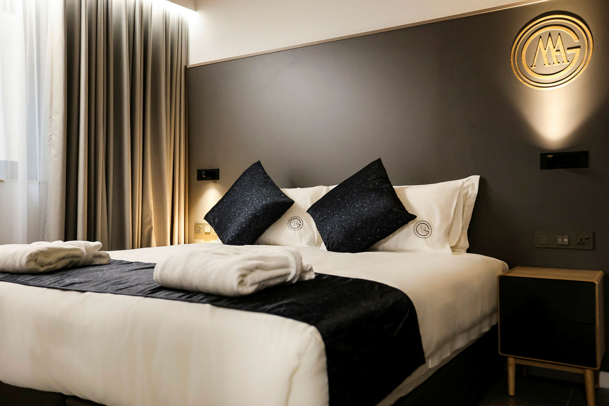 Et moderne hotelværelse med en dobbeltseng opredt med hvidt sengetøj og mørke dekorative puder. To sammenlagte badekåber ligger på sengen. En væg i rummet er mørkegrå med et gyldent dekorativt element. (AI-genereret tekst)