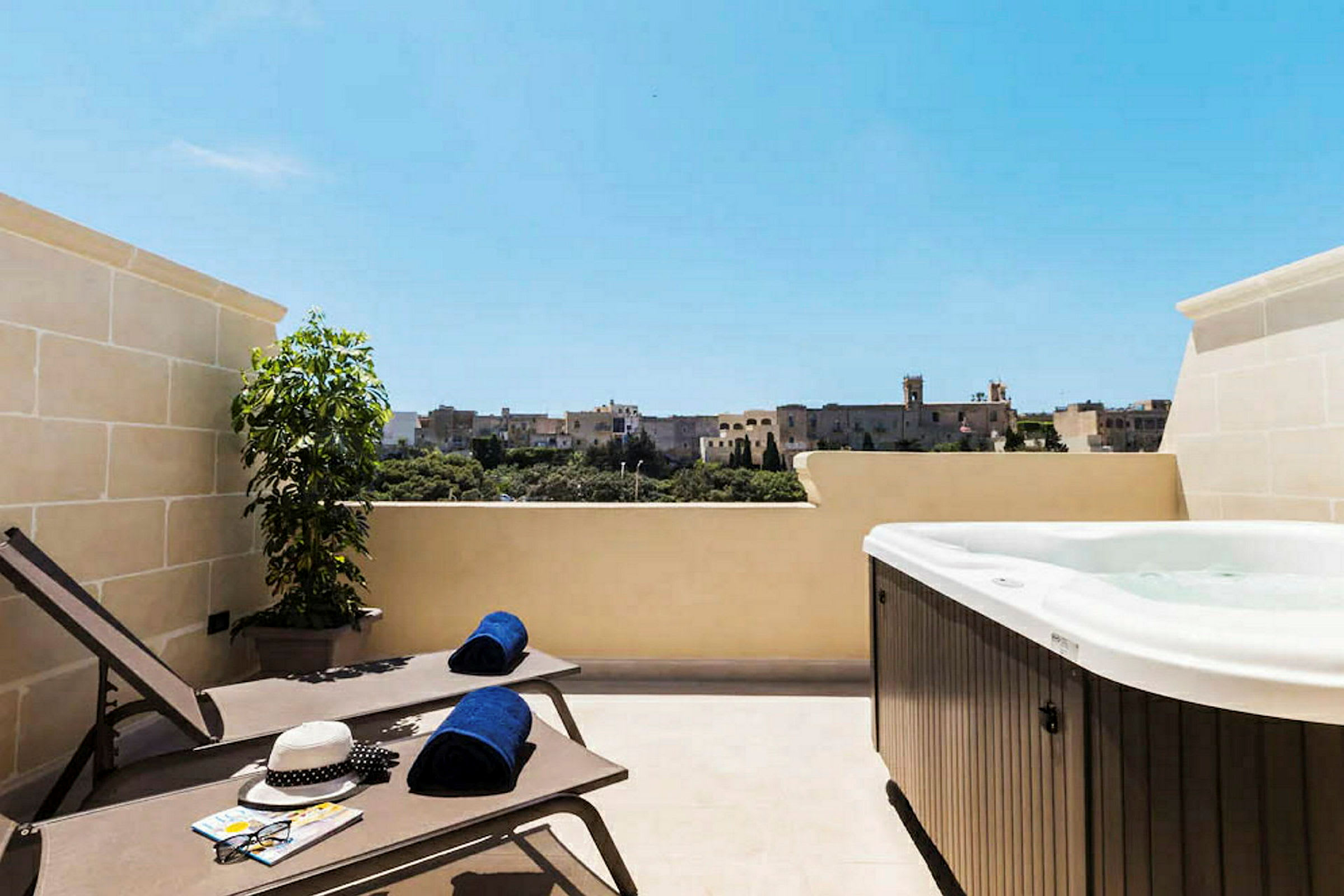 Et solrigt hotelværelse med en udendørs terrasse, liggestole, håndklæder og et spabad, med udsigt over en historisk bygning. (AI-genereret tekst)