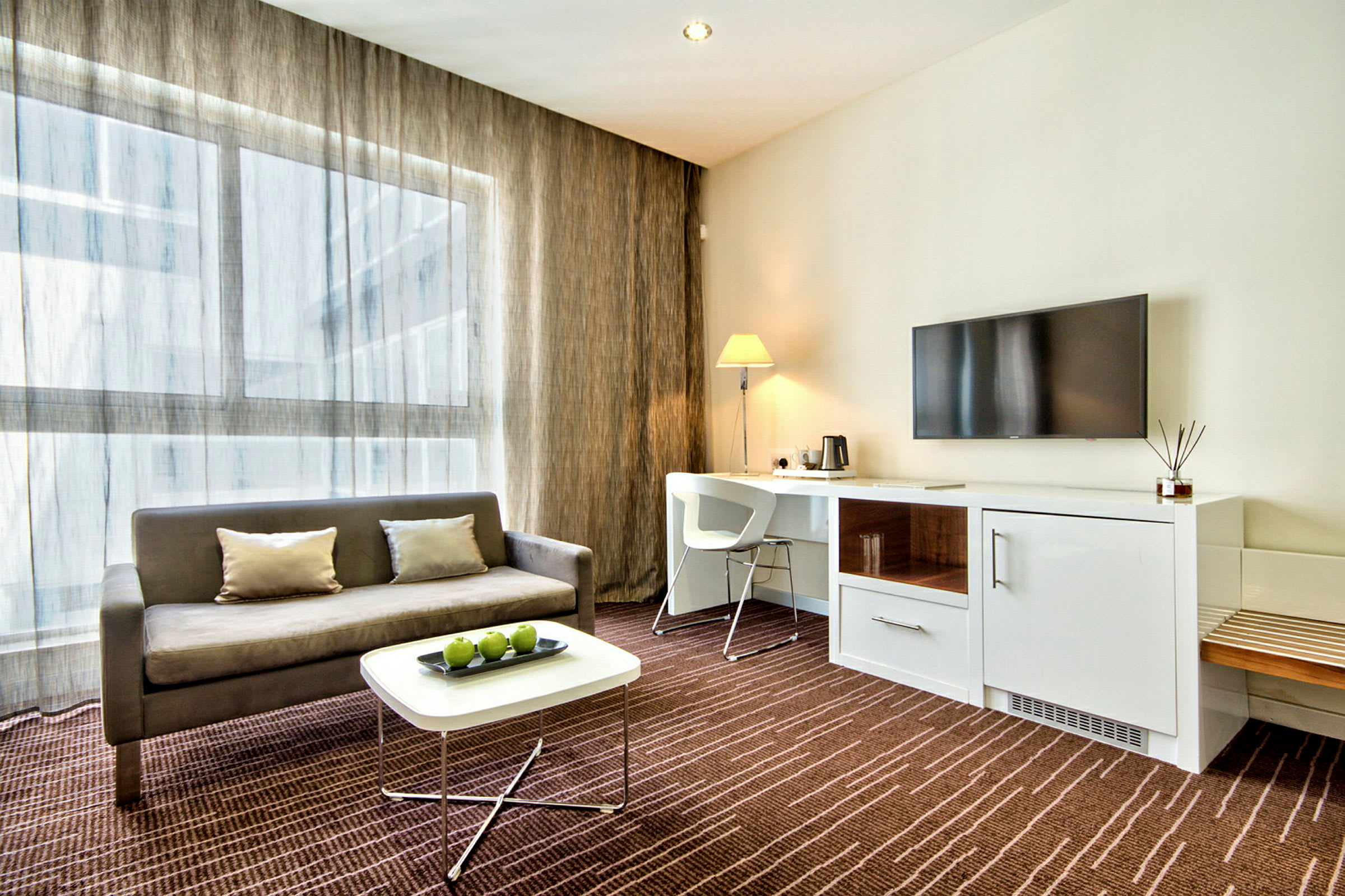 Et moderne hotelværelse med en sofa, et sofabord med grønne æbler, et arbejdsområde med stol og lampe samt et vægmonteret TV. (AI-genereret tekst)