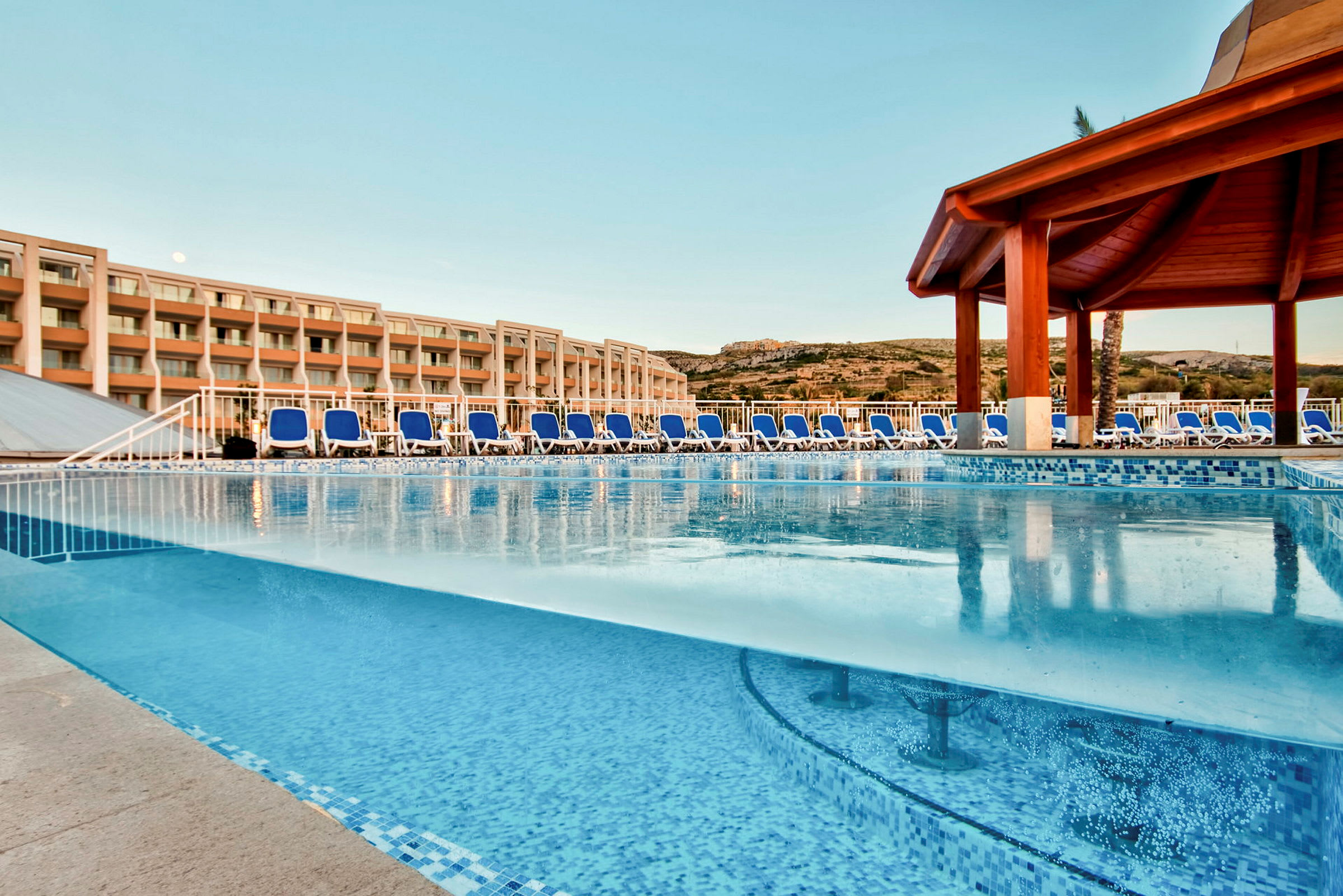 Udendørs swimmingpool med liggestole og hotelbygning i baggrunden. (AI-genereret tekst)