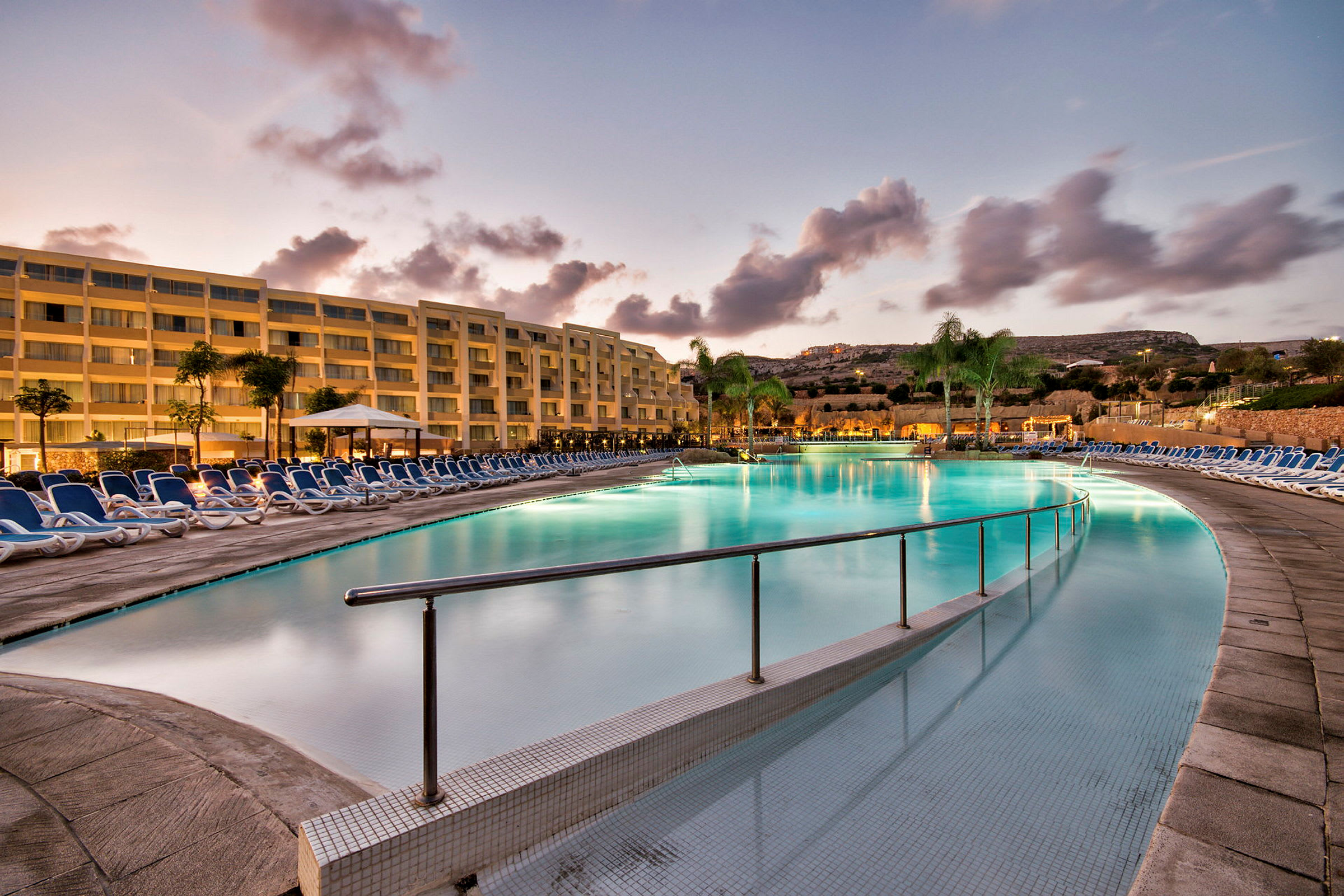 En stor udendørs swimmingpool med solsenge og en boligblok i baggrunden. (AI-genereret tekst)