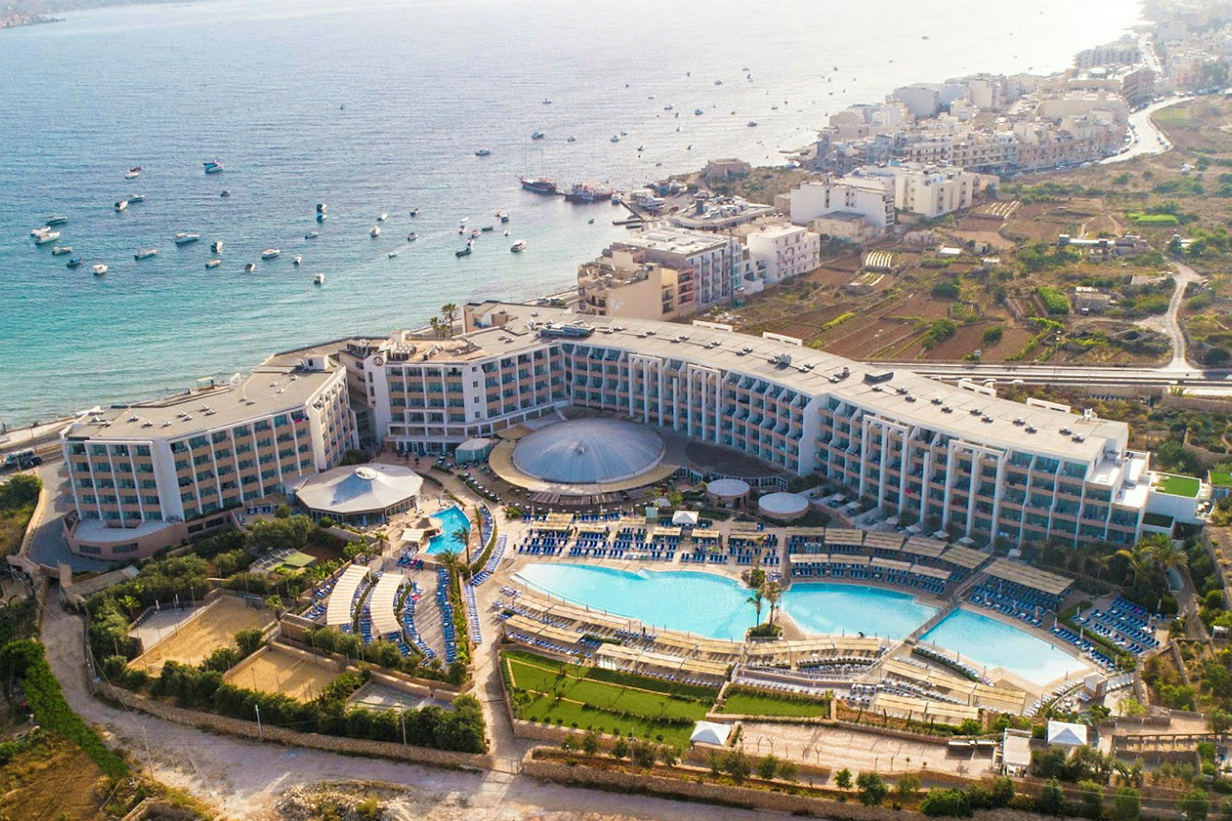 Luftfoto af et hotelkompleks med pools, grønne områder og havudsigt. (AI-genereret tekst)