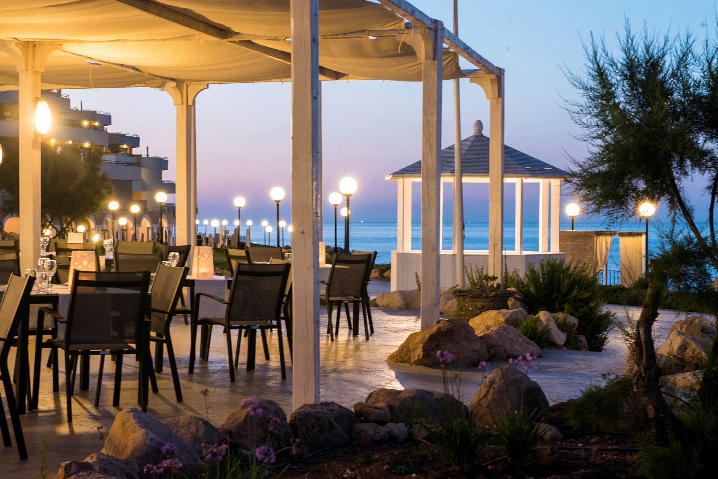 Et oplyst udendørsområde med borde og stole under et tag tæt på havet. Lamper skaber en behagelig atmosfære, og en hvid pavillon ses i baggrunden. (AI-genereret tekst)
