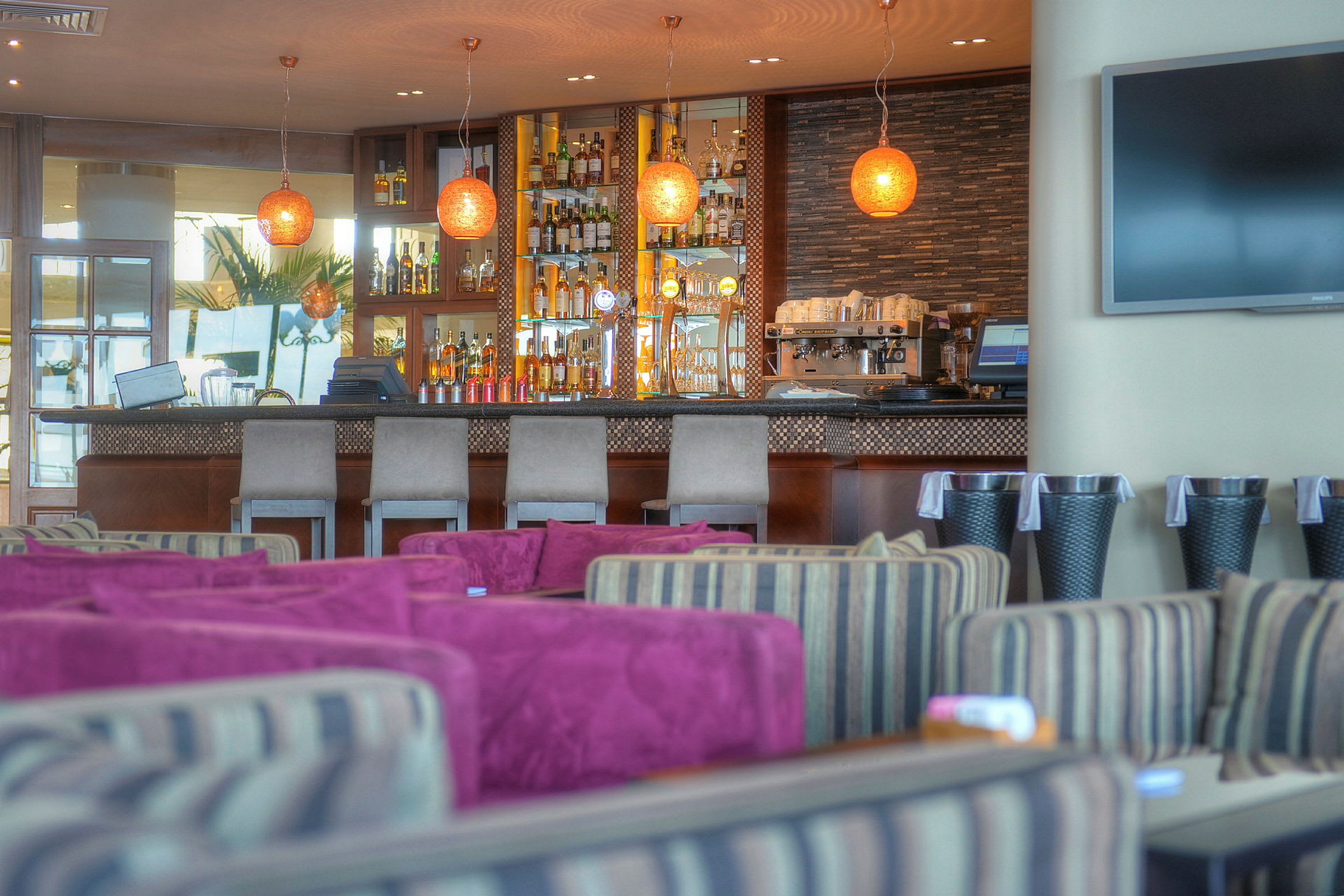 Et interiørbillede af en elegant bar med rækker af drikkevarer, pæne barstole og moderne lamper. I forgrunden er der siddepladser med farverige puder. (AI-genereret tekst)