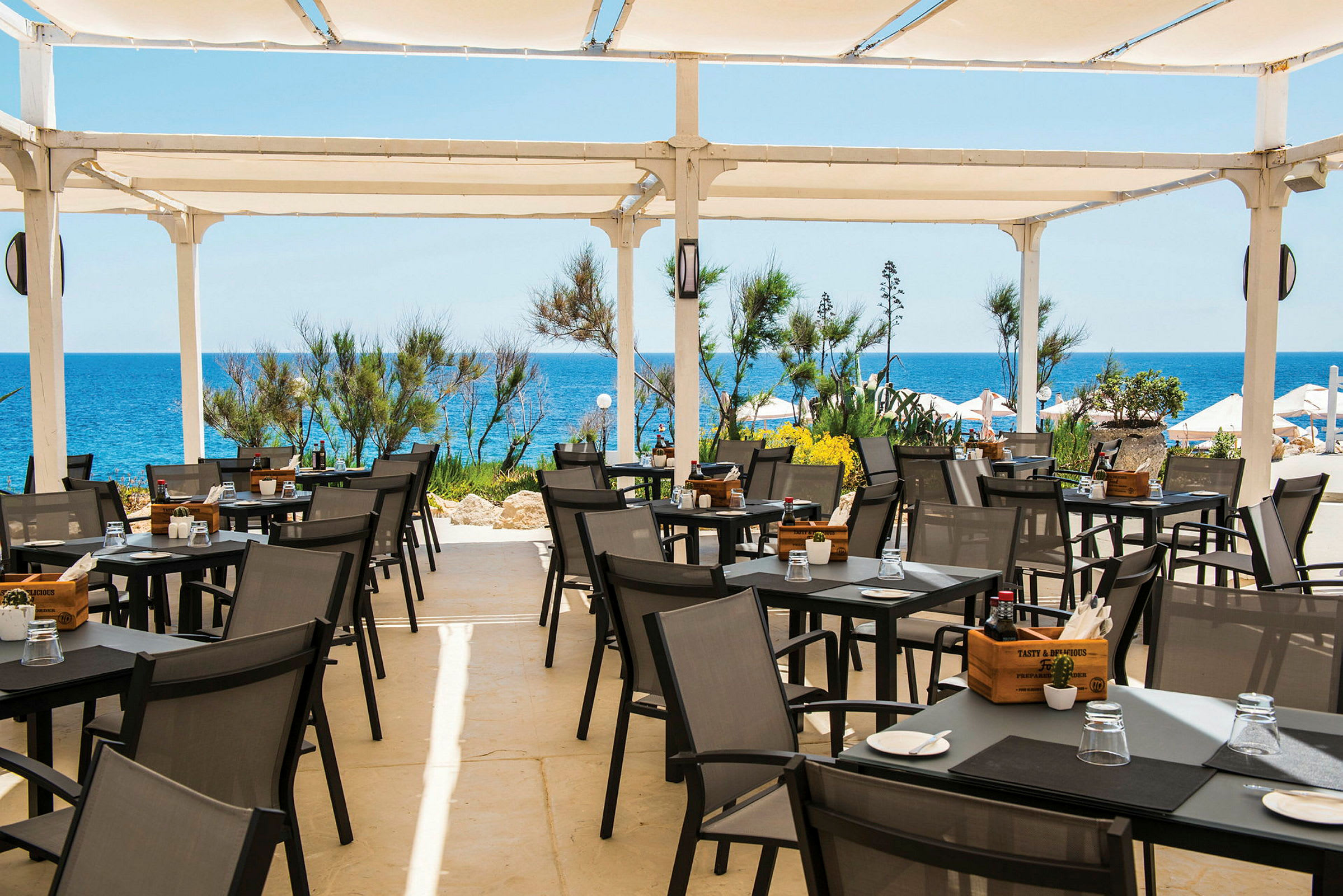 En terrasse med udendørsservering, borde og stole under et hvidt tag, med havet og vegetation i baggrunden. (AI-genereret tekst)