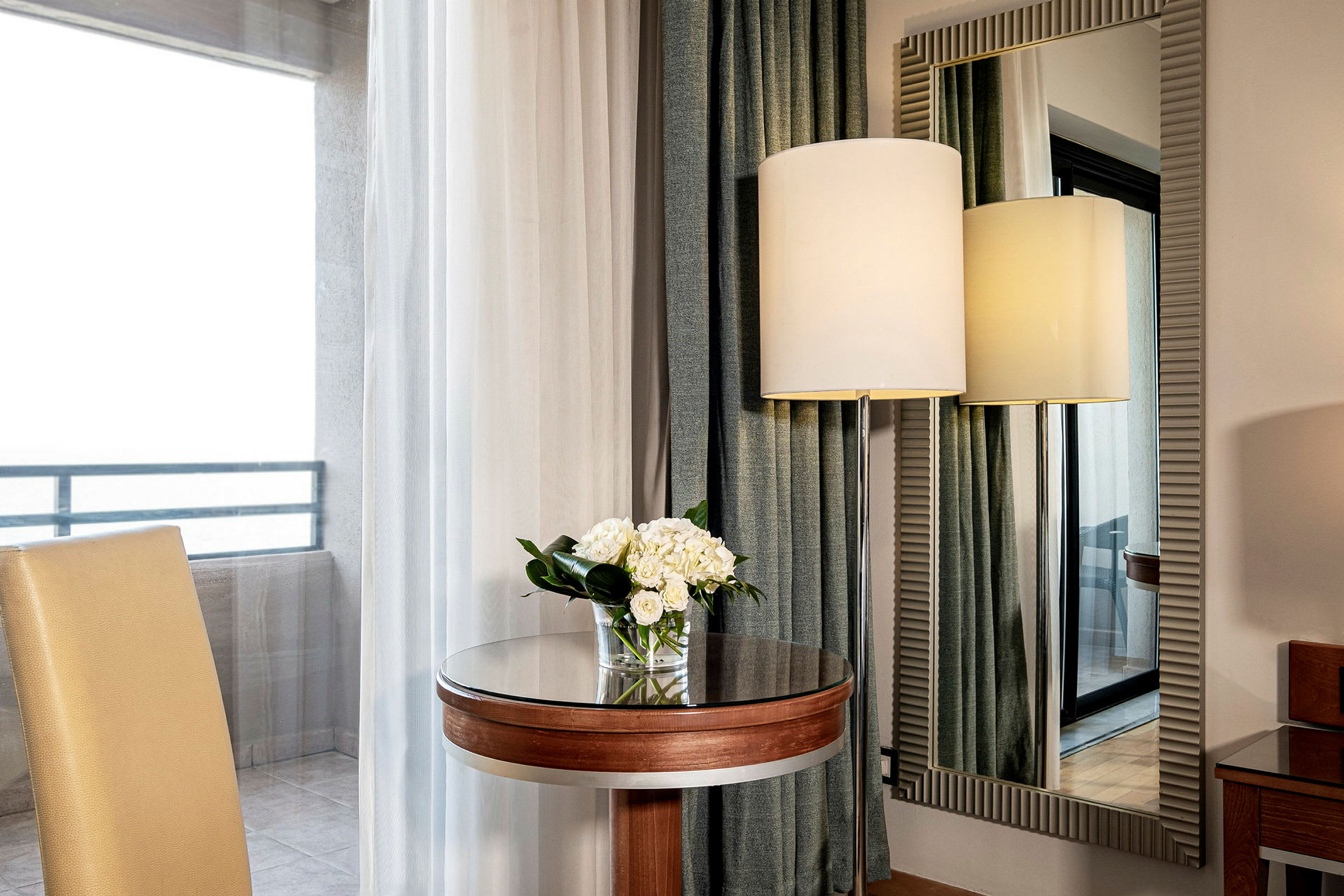 Et elegant hotelområde med en stol, et lille rundt bord med en blomsterbuket, et stort spejl og en gulvlampe set gennem et vindue. (AI-genereret tekst)