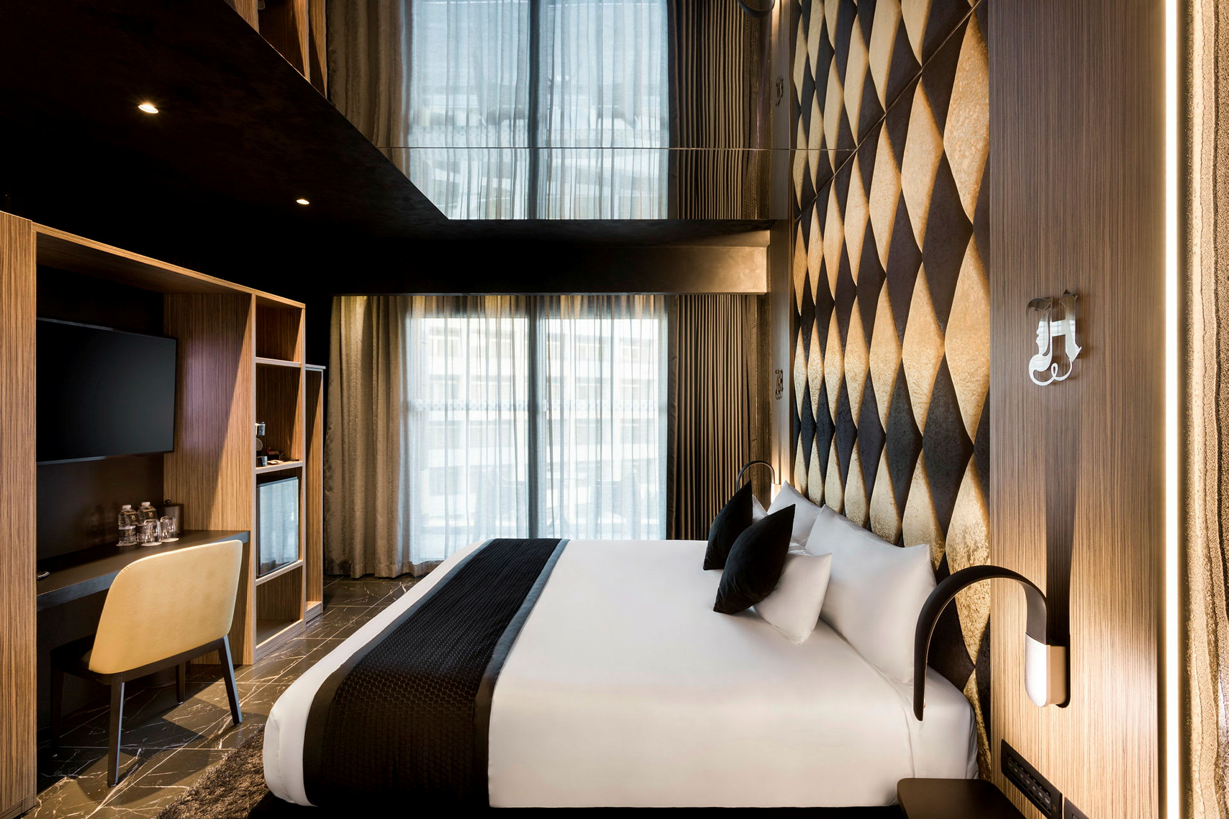 Et moderne hotelværelse med en bred seng dækket med hvidt sengetøj og sorte puder. Væggene har et dekorativt diamantmønster, og ved siden af sengen er der et lille skrivebord med en stol. (AI-genereret tekst)