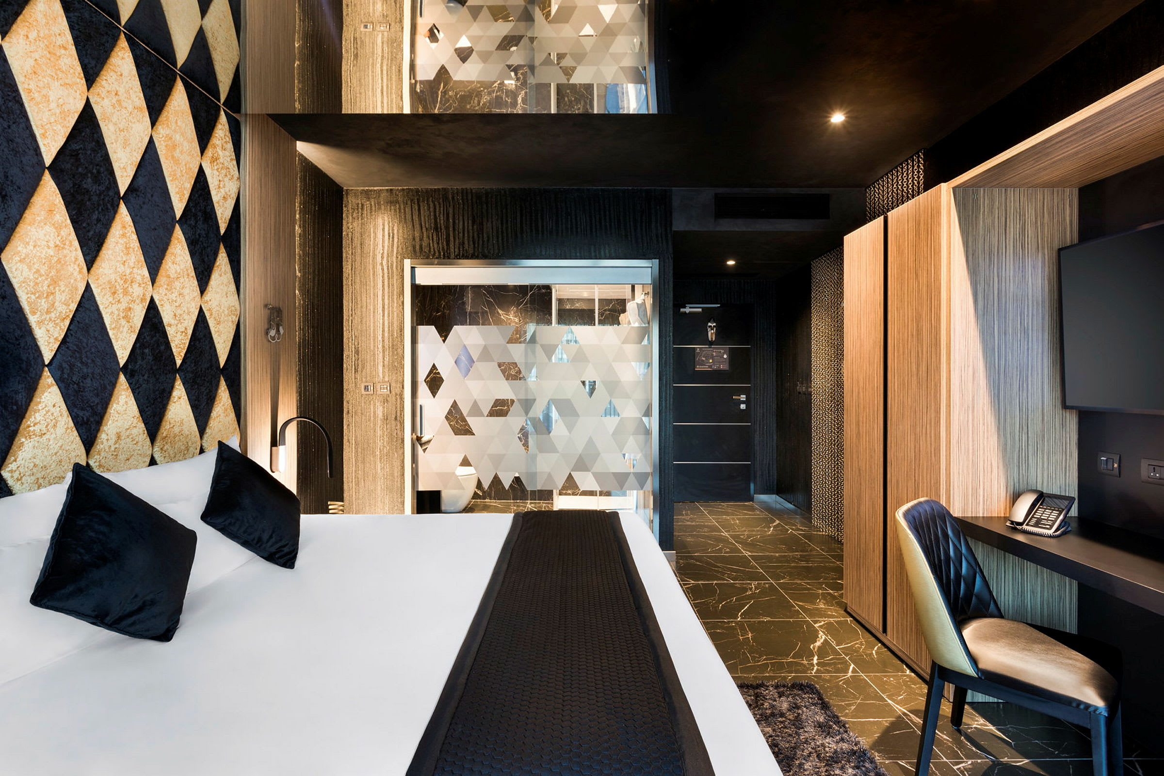 Hotelværelse med moderne design, seng med hvide lagner og sorte puder samt arbejdsområde med stol, skrivebord og TV. (AI-genereret tekst)