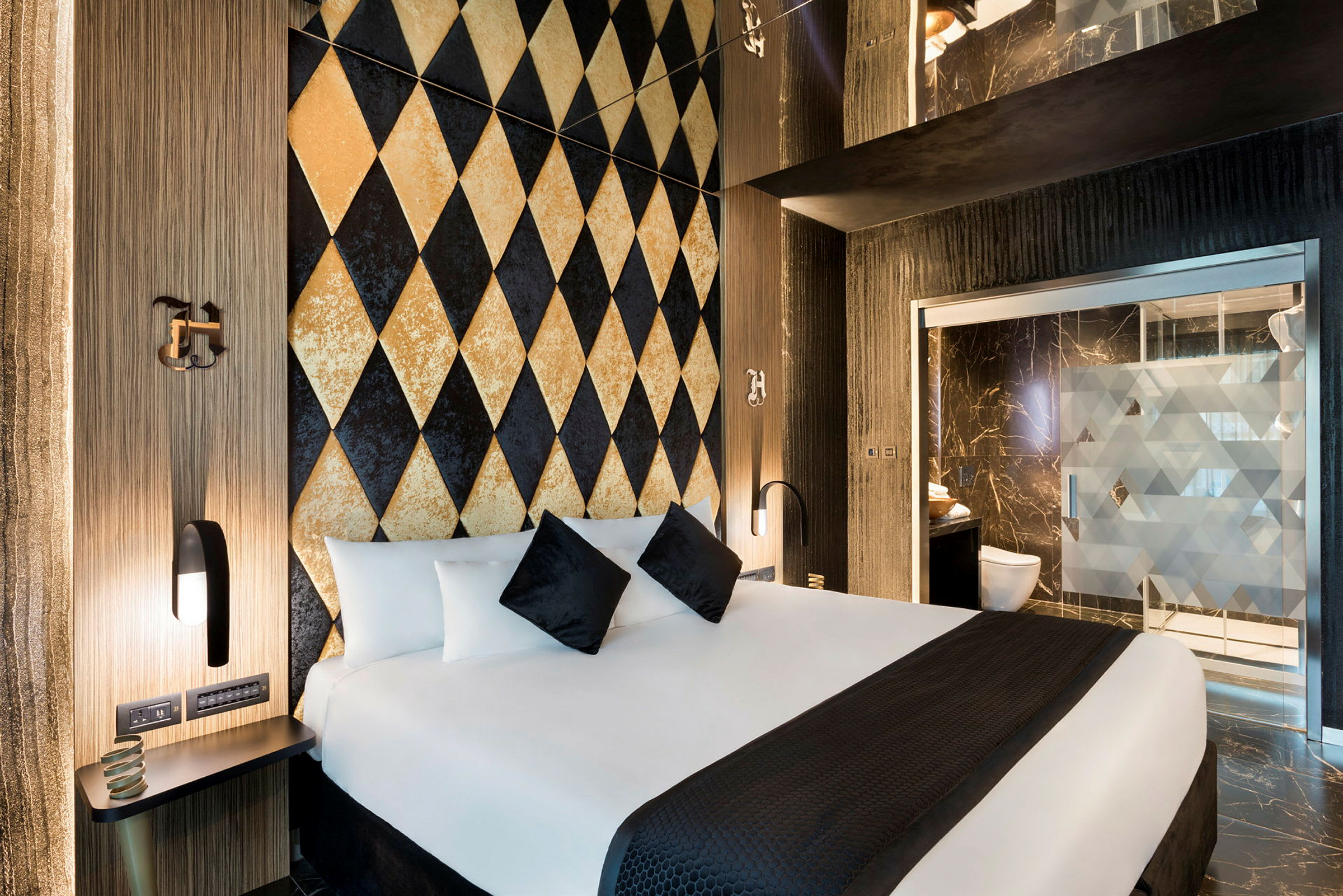 Et moderne hotelværelse med en dekorationsvæg i harlekinmønster og en stor seng med hvide og sorte puder. Badeværelset ses i baggrunden. (AI-genereret tekst)