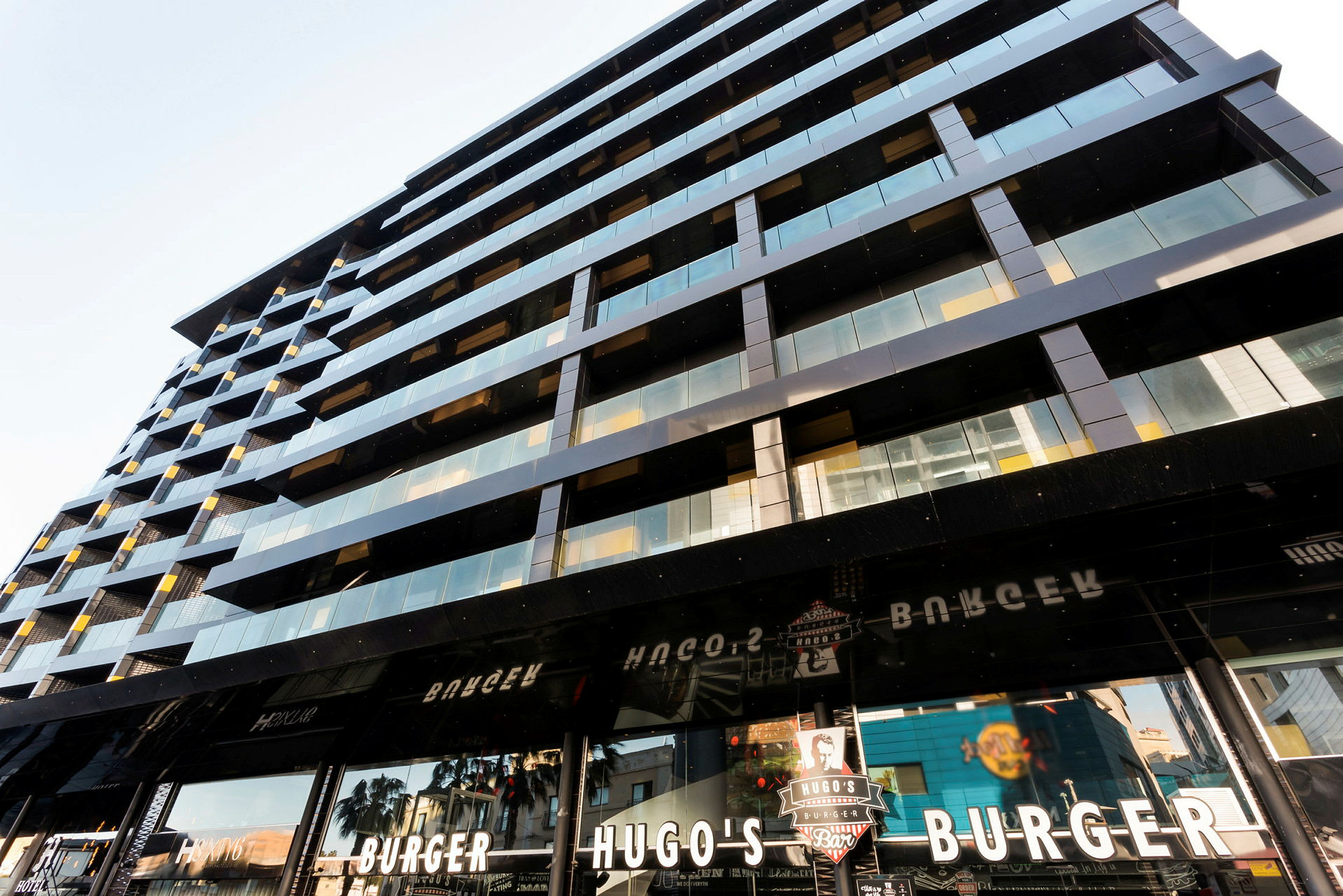 En moderne bygning med glas- og metalfacade samt skilte for en burgerrestaurant kaldet Hugo's Burger. (AI-genereret tekst)