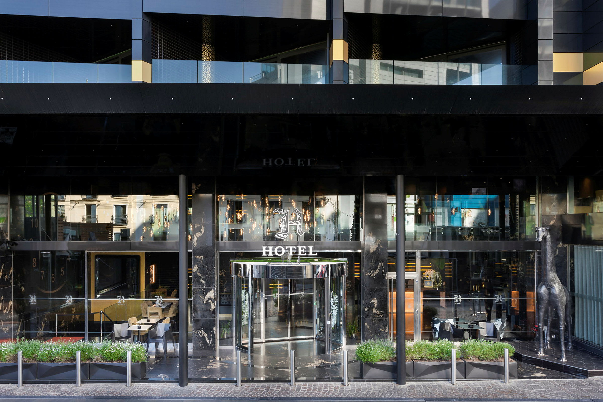 Forsiden af et hotel med glasdøre og en dekorativ sølvfarvet girafstatue ved siden af indgangen. (AI-genereret tekst)