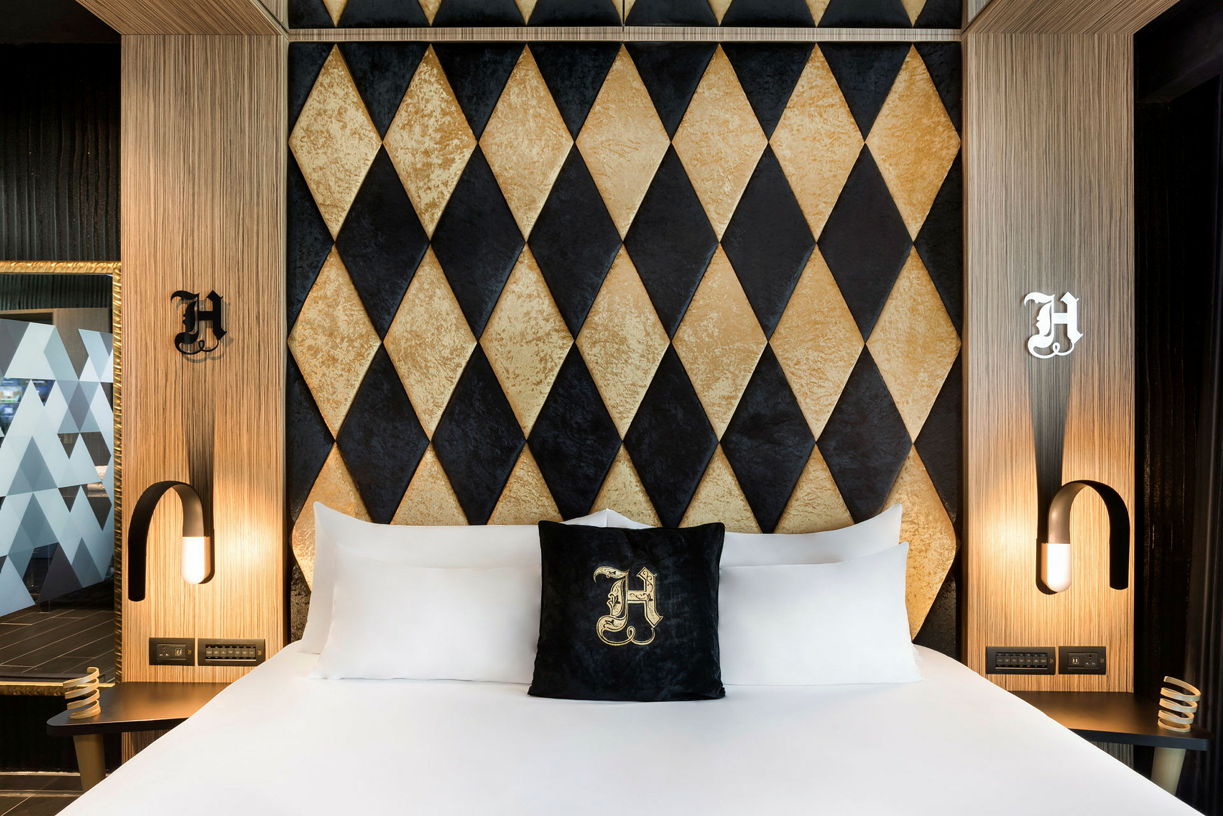 Et hotelværelse med en seng med hvide lagner og en dekorativ pude med bogstav. Bag sengen er en væg med diamantmønster i sort og guld. (AI-genereret tekst)