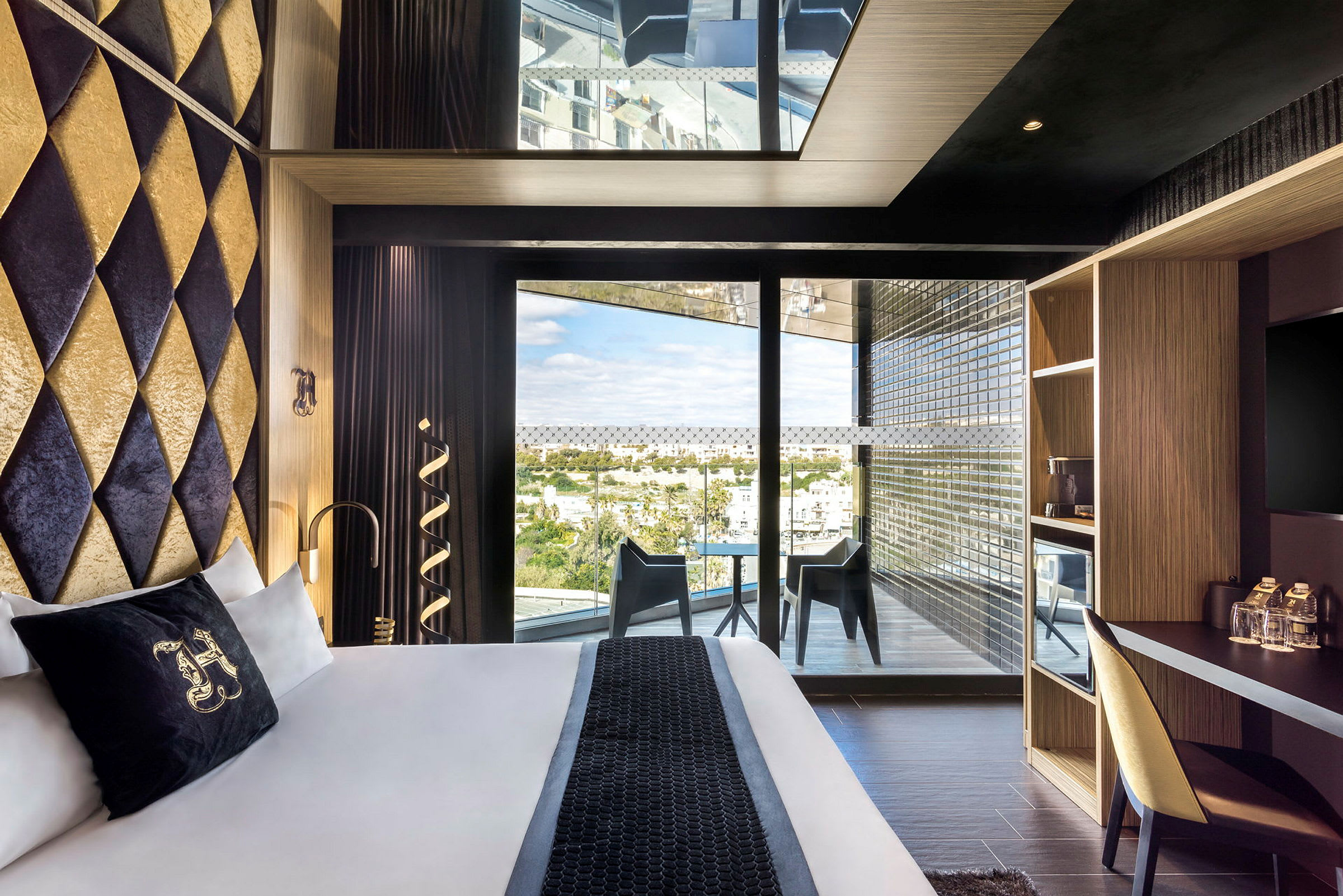 Interiør af et moderne hotelværelse med dobbeltseng, skrivebord, tv og en balkon med siddepladser og udsigt over byen. (AI-genereret tekst)