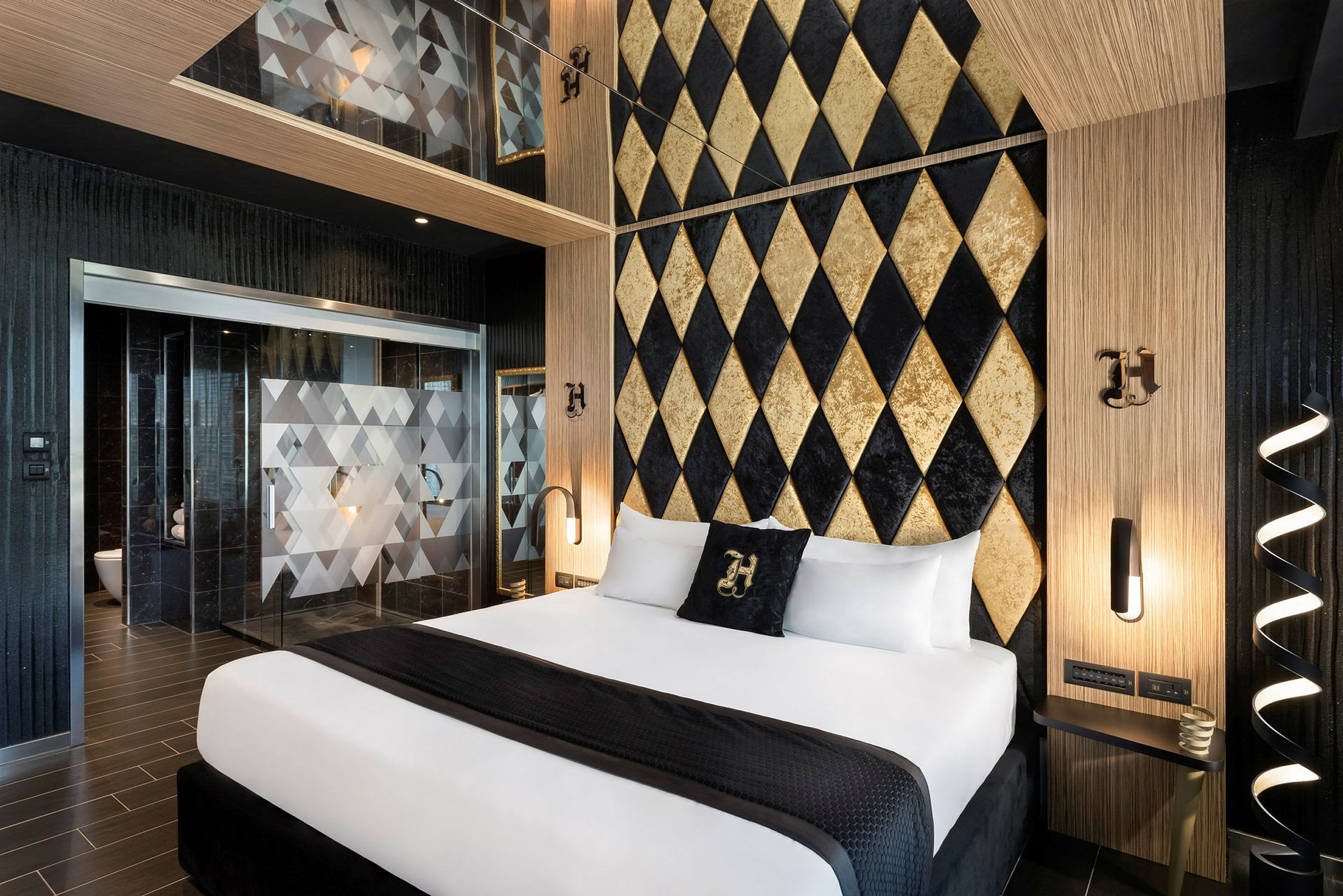 Et hotelværelse med elegant design. En dobbeltseng med hvide lagner og et sort tæppe er centralt placeret. Baggrunden er en væg med guldfarvede og sorte diamantmønstre. Der er adskilte glasdøre med geometriske detaljer, der fører til et badeværelse. (AI-genereret tekst)