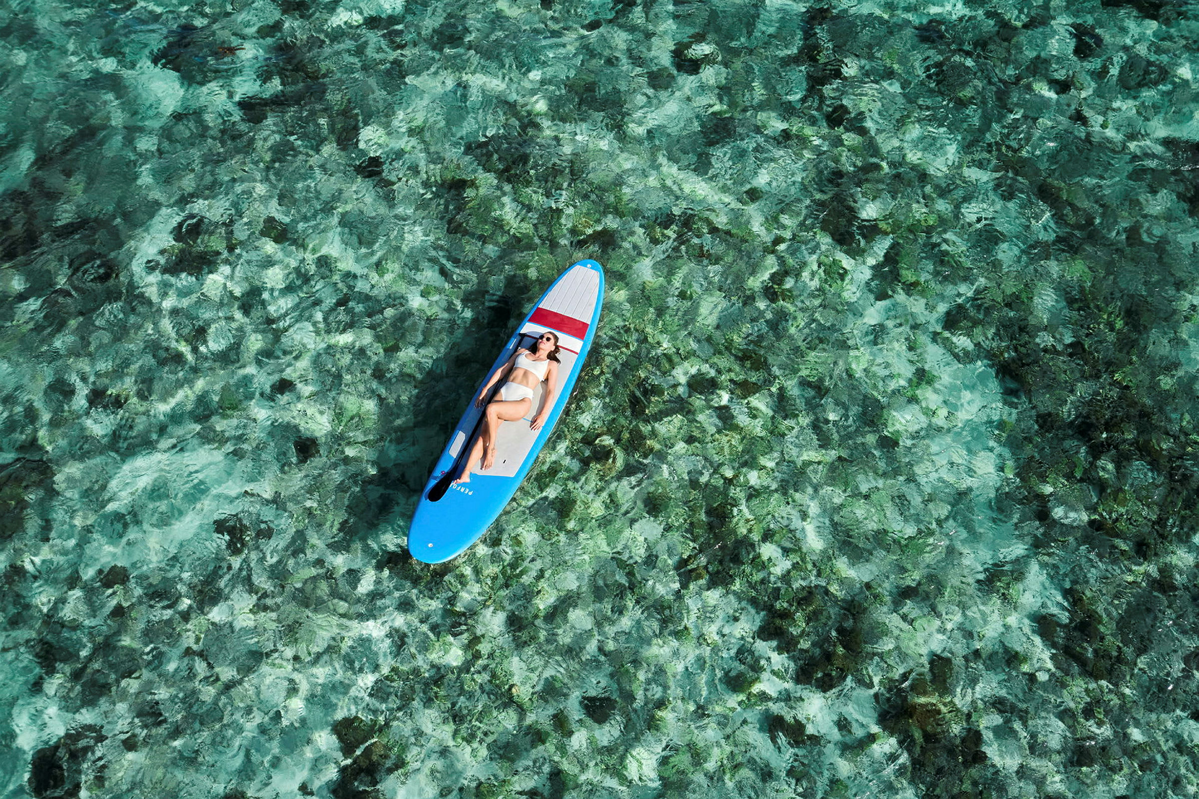 En person ligger afslappet på et paddleboard over klart turkis vand. (AI-genereret tekst)