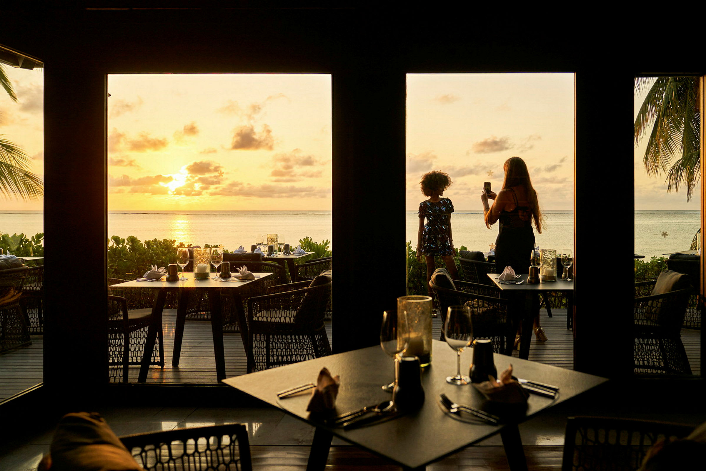 Billedet viser en restaurant med udsigt over en strand ved solnedgang, med borde anbragt både indenfor og udenfor, samt palmer og havet i baggrunden. (AI-genereret tekst)