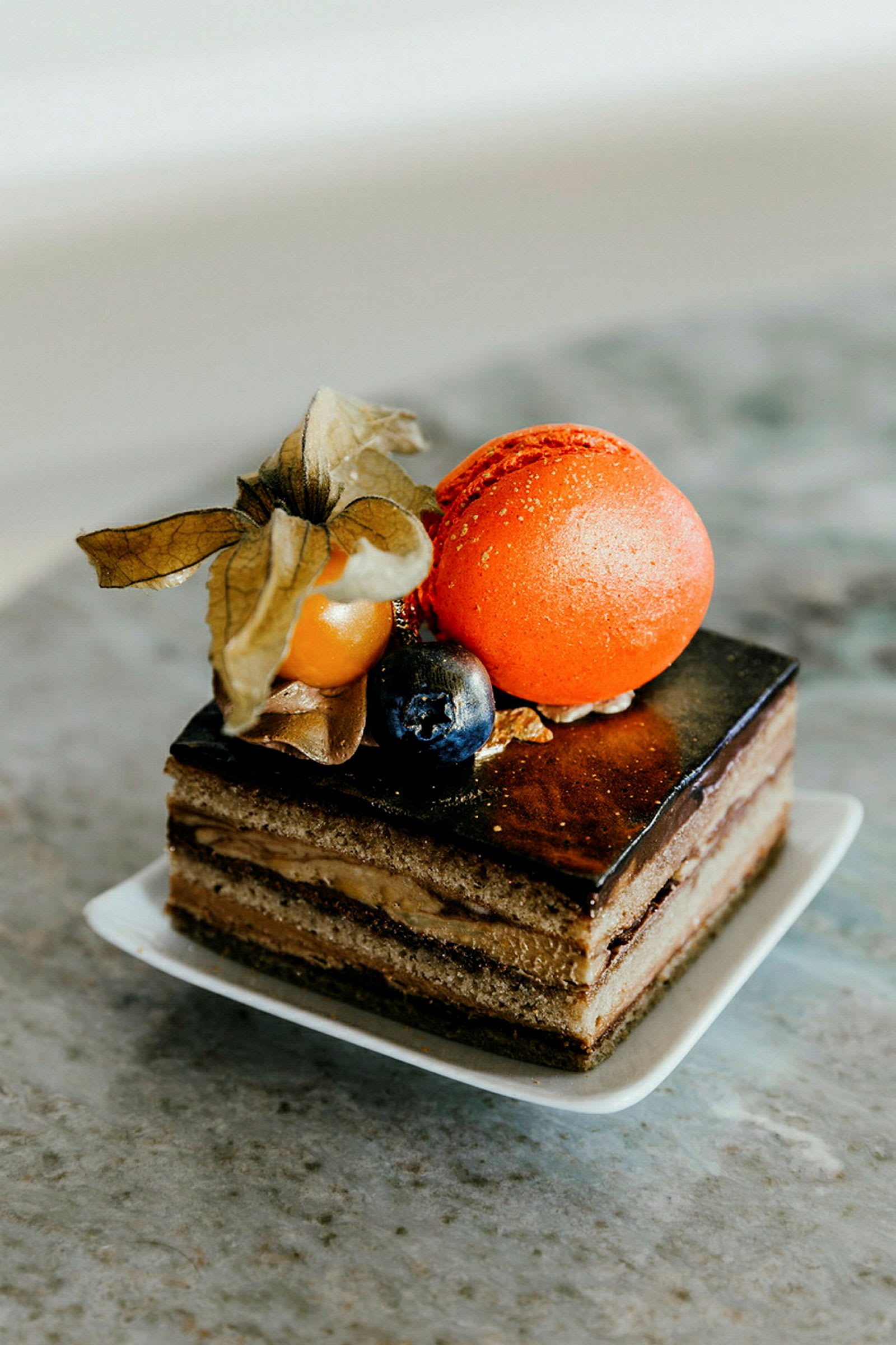 Nærbillede af en elegant dessert med chokoladeglasur, lag af creme, dekoreret med en orange makron, en physalis og et blåbær. (AI-genereret tekst)