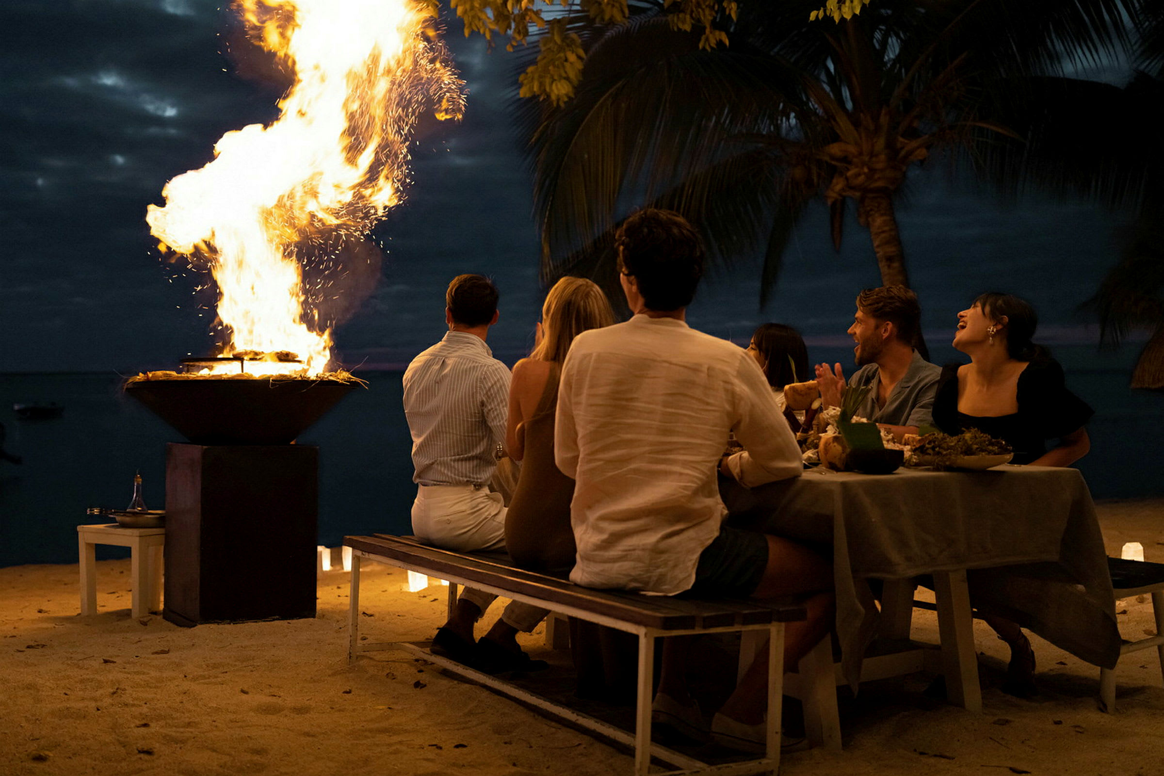 En gruppe mennesker sidder ved et dækket bord på stranden tæt på en ildskål med flammer om aftenen. (AI-genereret tekst)