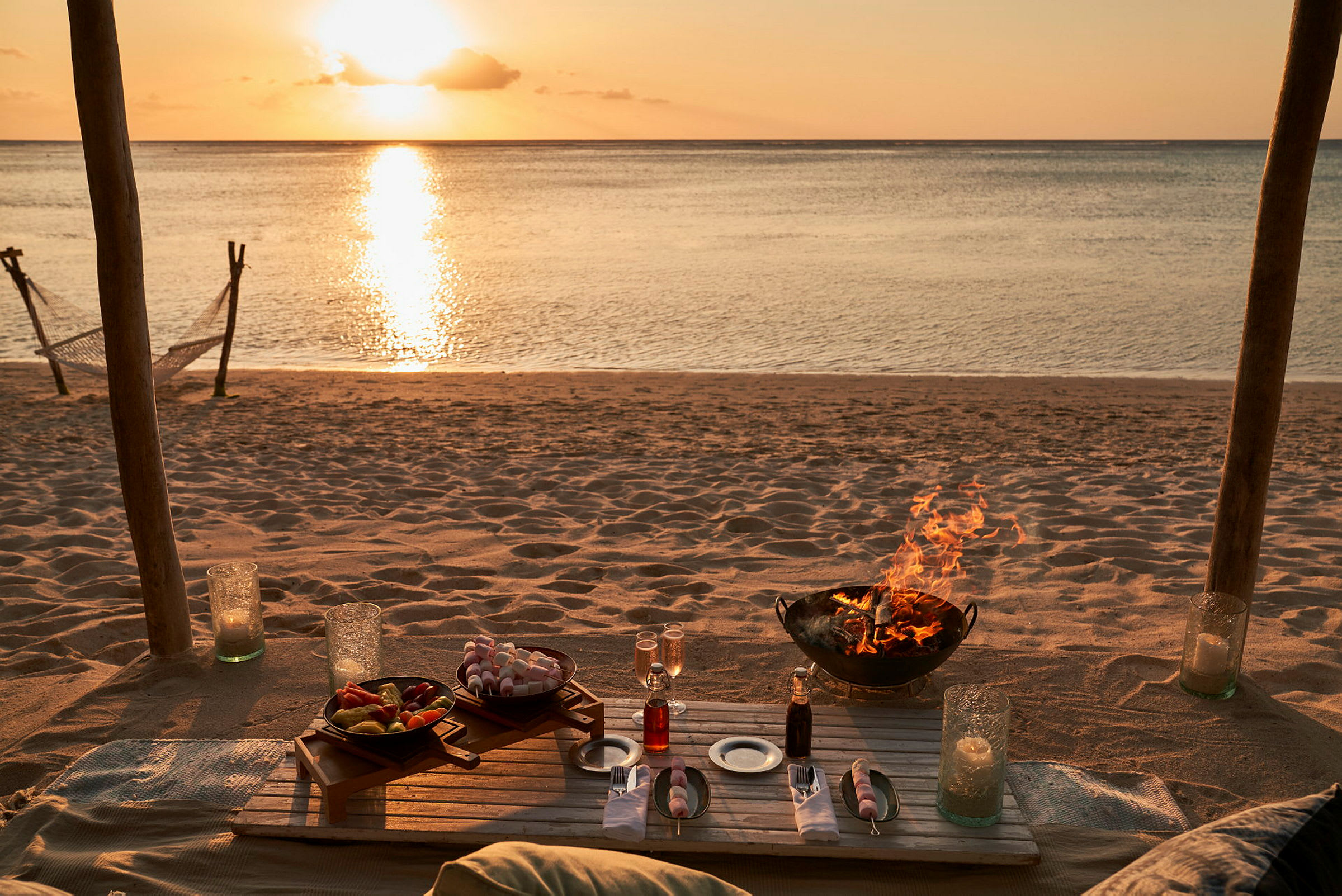 Dækket bord på en strand med en solnedgang i baggrunden og et bål i en ildsted. (AI-genereret tekst)