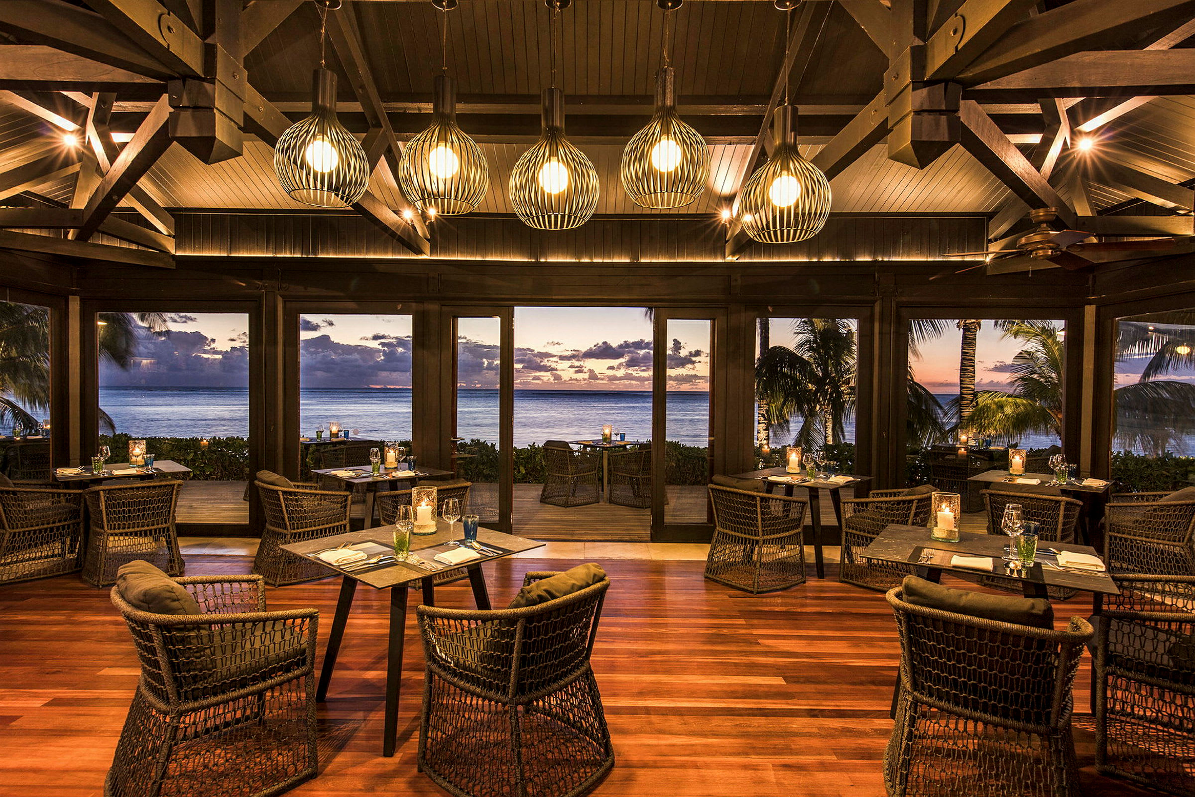 En elegant restaurant med træindretning og moderne stole. Vinduerne giver udsigt over havet med en solnedgang i det fjerne. (AI-genereret tekst)