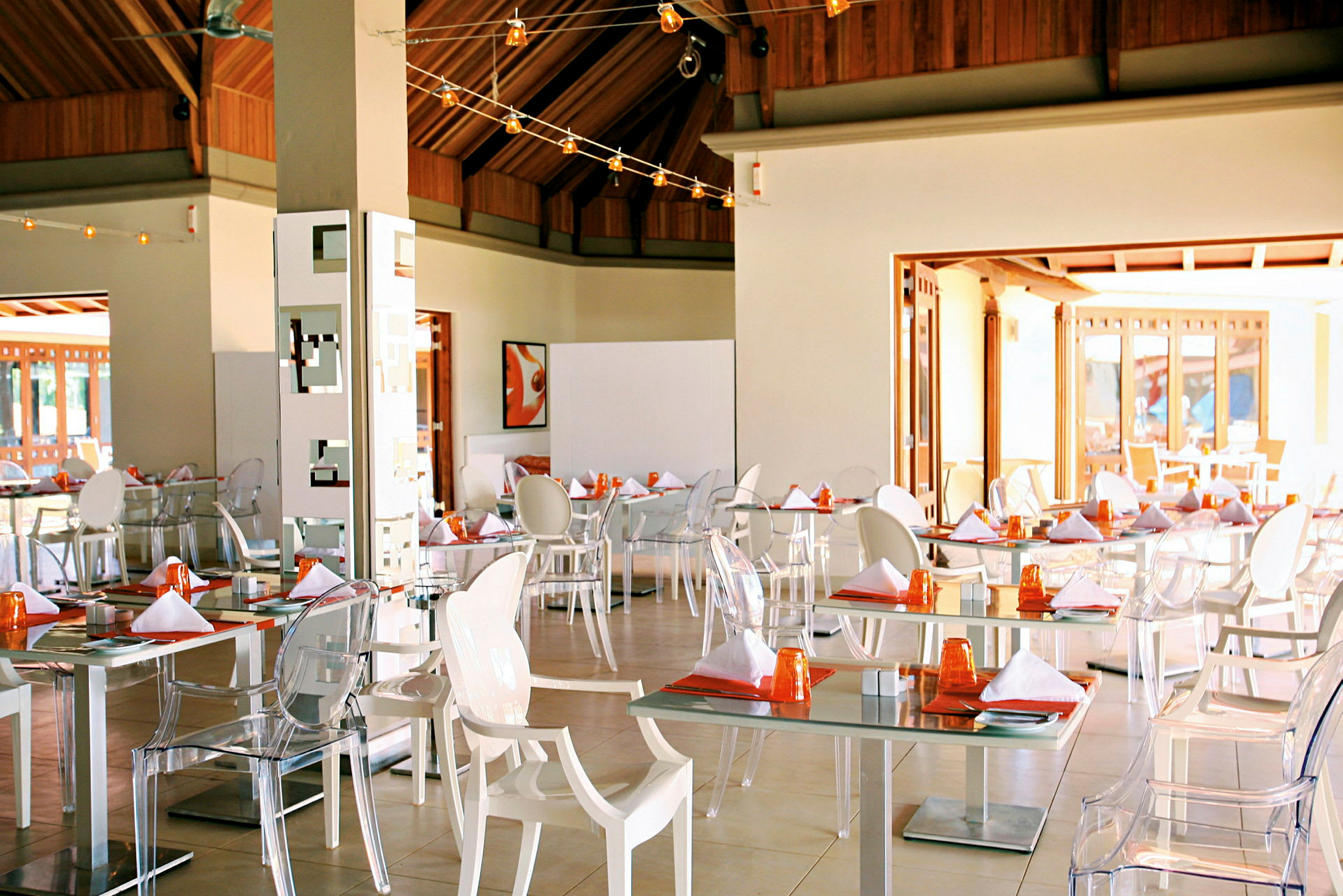 Et moderne indrettet restaurantområde med hvide borde, gennemsigtige og hvide stole samt borddækning med tallerkener og orange glas. (AI-genereret tekst)