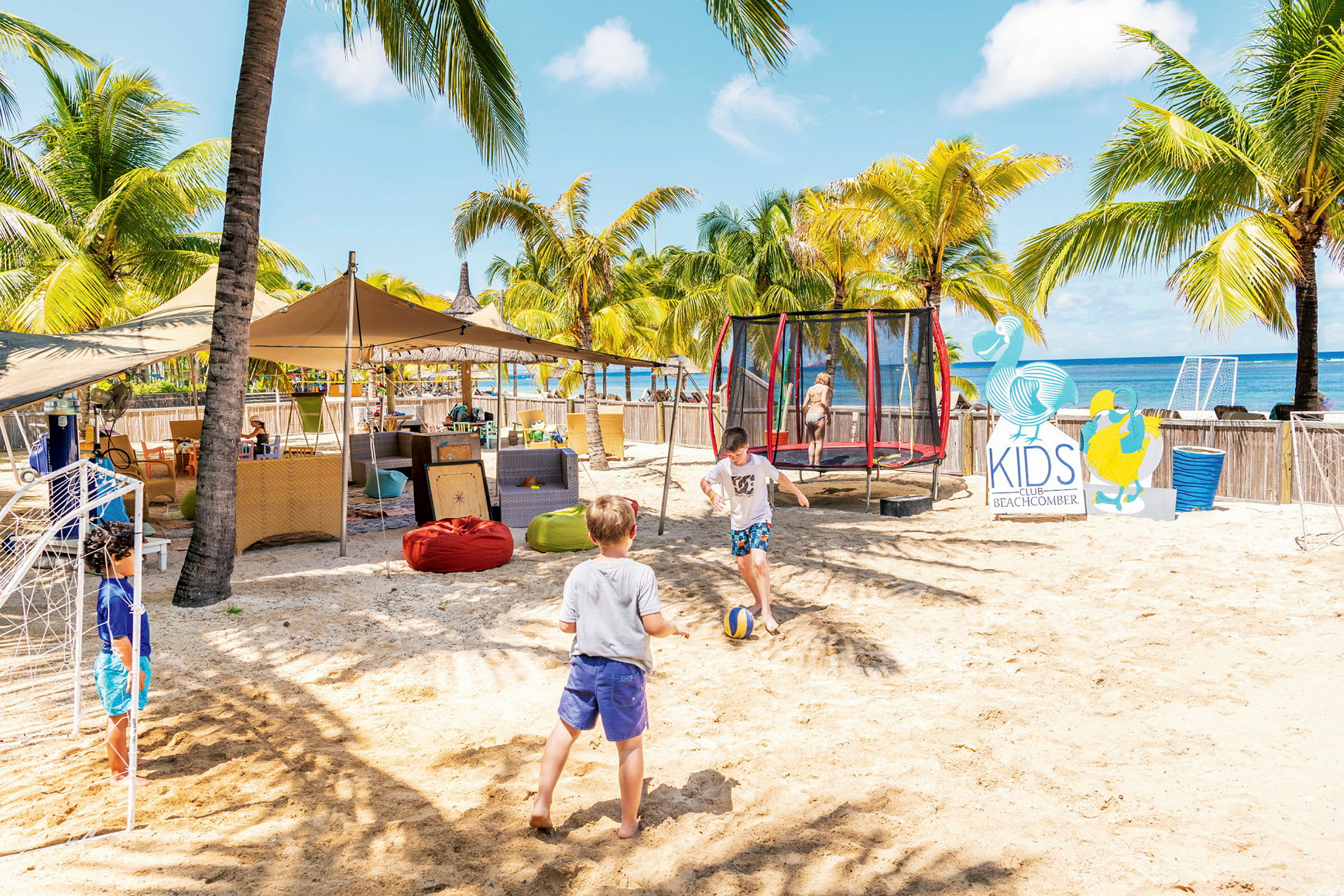 Børn leger på en sandstrand ved siden af en trampolin og et skilt for Kids Club. (AI-genereret tekst)