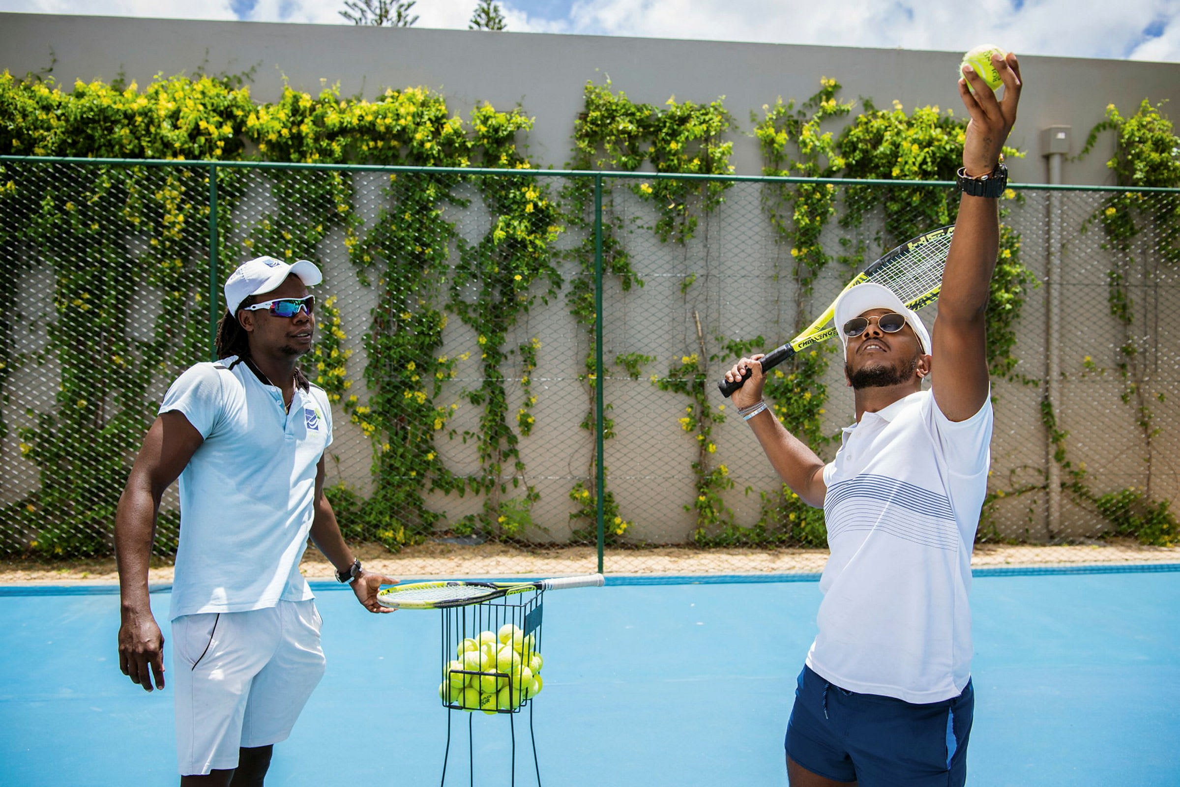 To personer spiller tennis på en udendørs tennisbane med en beholder fyldt med tennisbolde. (AI-genereret tekst)