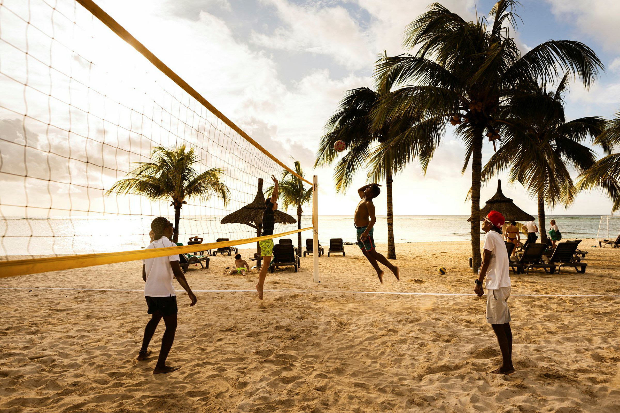 Fire personer spiller strandvolleyball på en sandstrand med liggestole og palmer i baggrunden. (AI-genereret tekst)