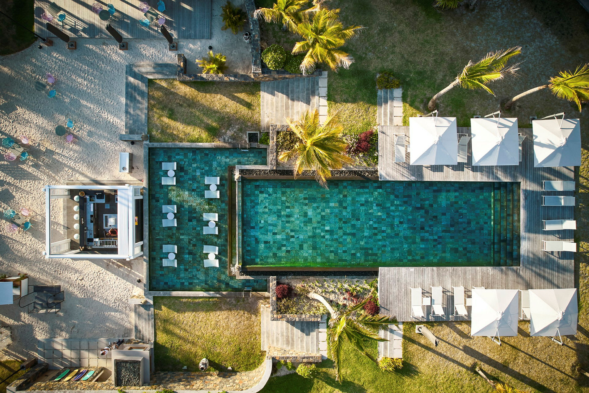 Luftfoto af en pool med liggestole og parasoller ved siden af samt omkringliggende grønne områder og bygninger. (AI-genereret tekst)
