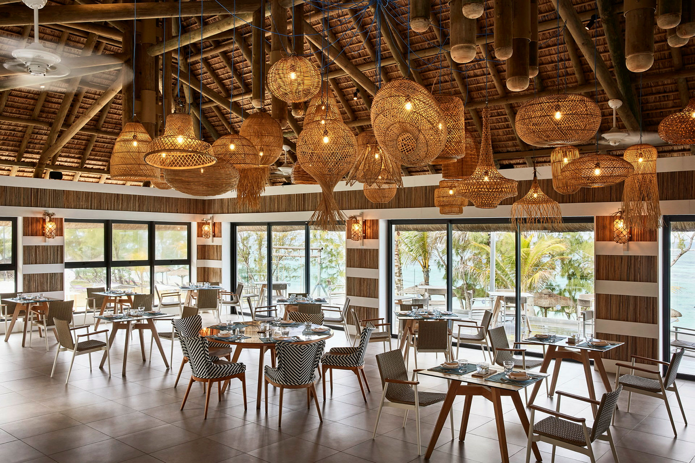 En restaurant med borde og stole, store vinduer med udsigt til tropiske omgivelser og dekorative loftslamper. (AI-genereret tekst)