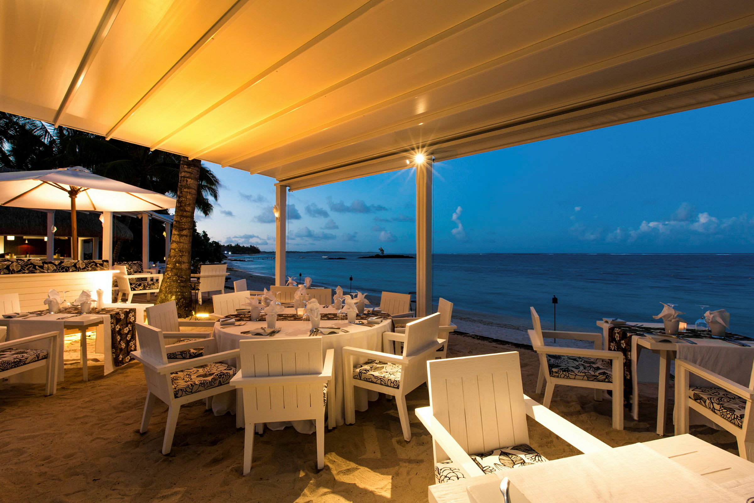 En restaurant med hvid møblering er beliggende ved stranden under en teltlignende tagstruktur. Havet kan ses i baggrunden med en klar blå himmel og diskrete skyer. (AI-genereret tekst)