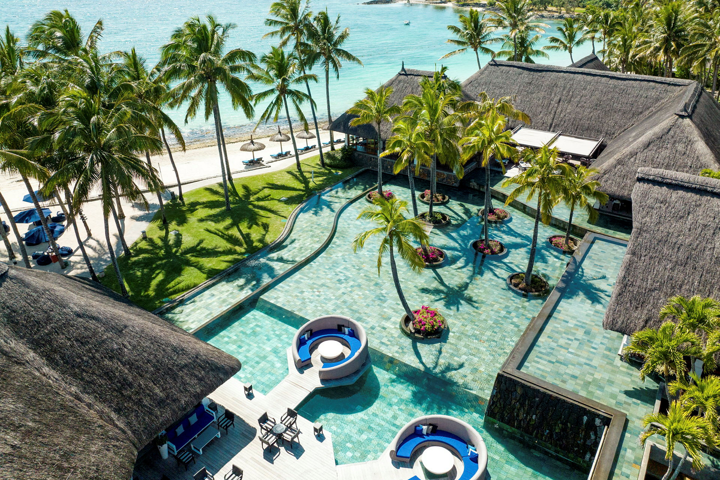 Flere huse med stråtag ved en pool med palmer og sandstrand i baggrunden. (AI-genereret tekst)