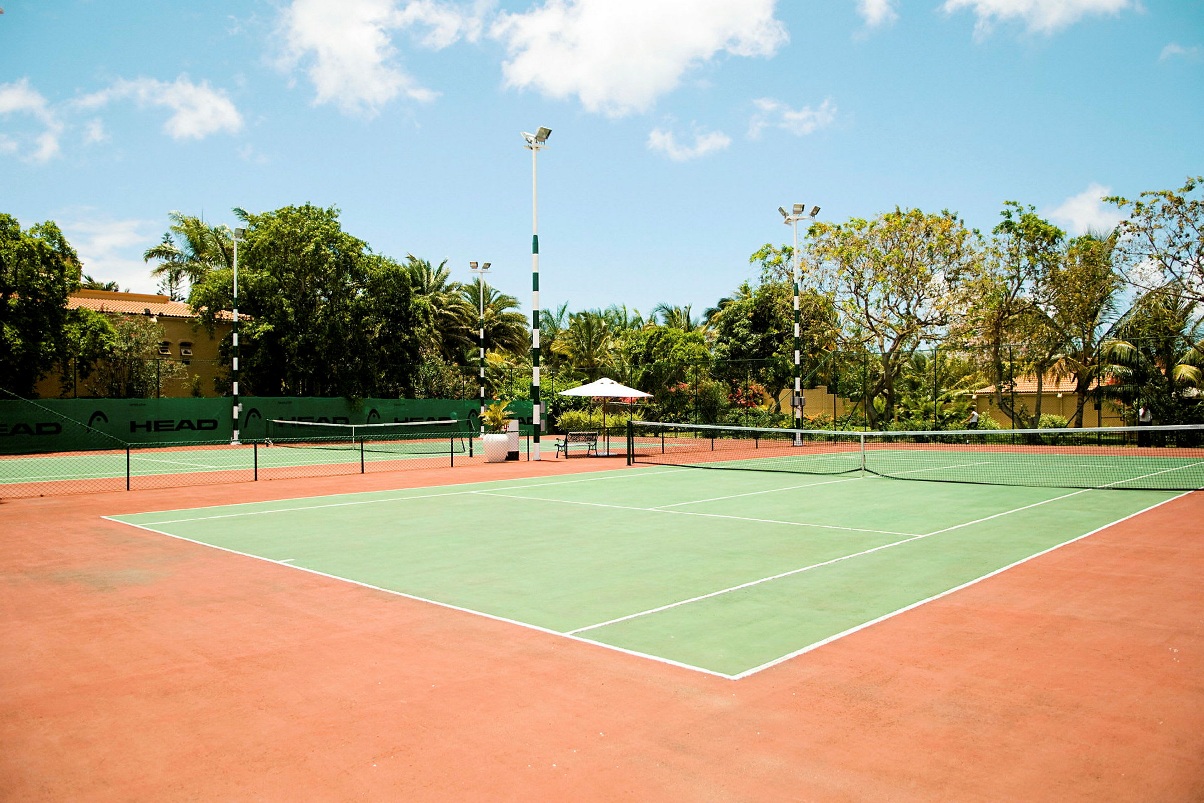 En tennisbane med grønt spilleområde og røde omkringliggende områder, omgivet af træer og bygninger. (AI-genereret tekst)