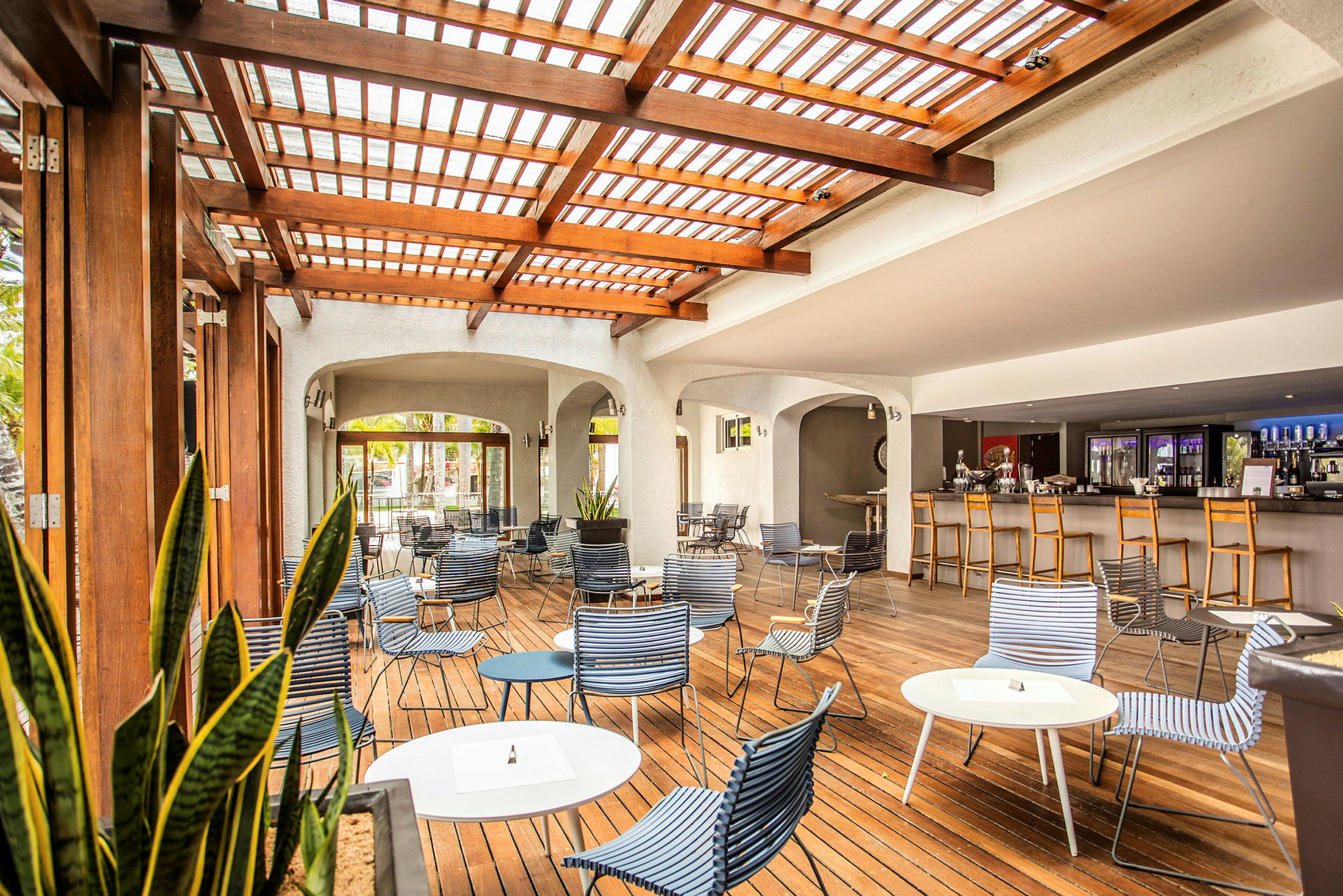 En udendørs lounge med træpergola, siddepladser og borde samt en bar i baggrunden. (AI-genereret tekst)