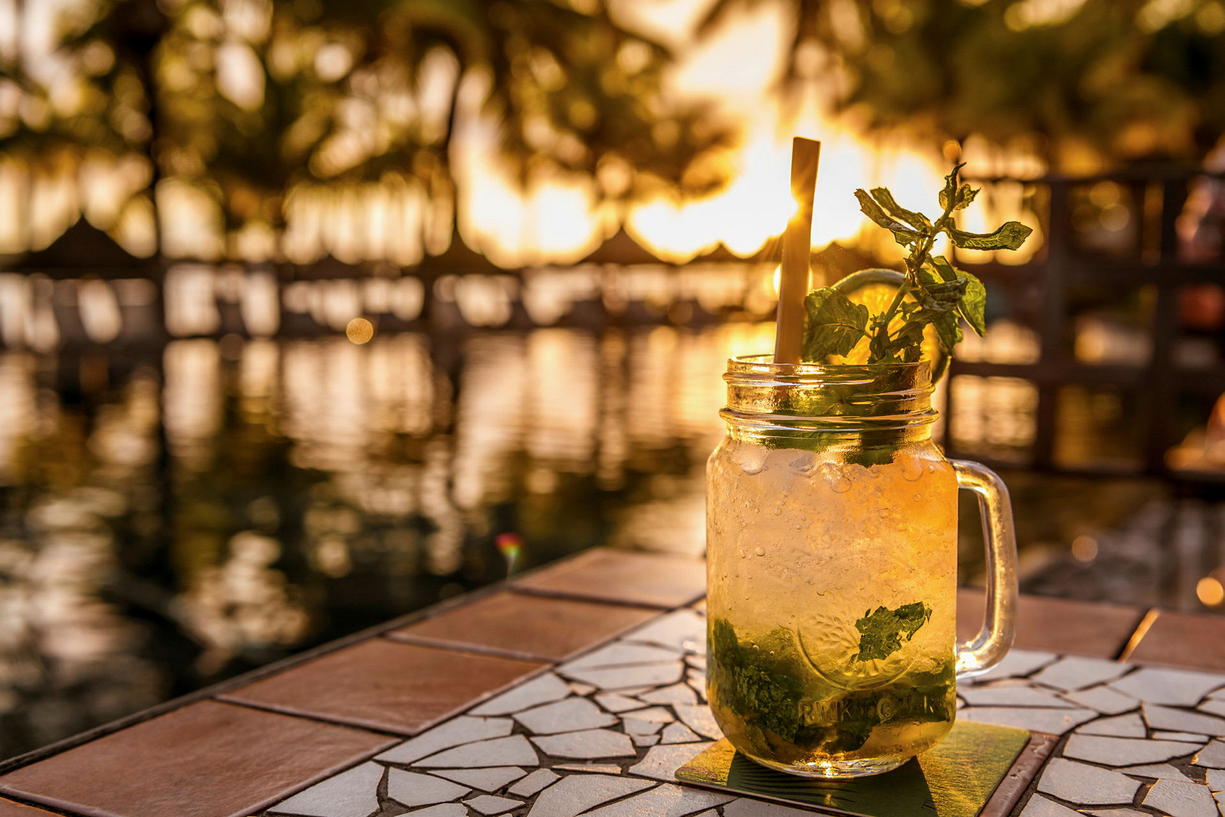 Et glas med en kold drink med mynte og citronskive står på et bord ved vandet i solnedgangen. (AI-genereret tekst)