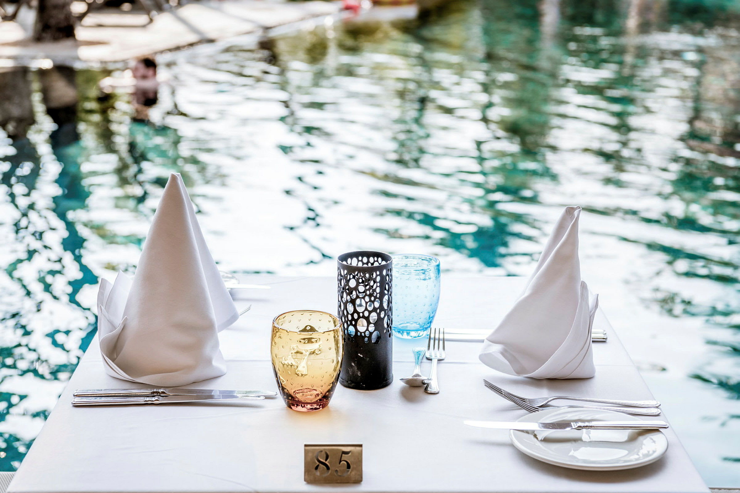 Et dækket bord med hvide servietter, glas og porcelæn ved siden af en pool. (AI-genereret tekst)