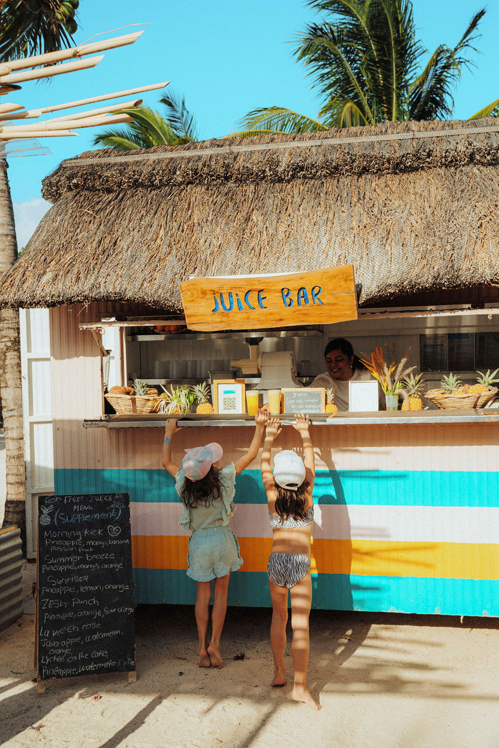 To personer står ved en juicebar med stråtag på en solrig plads. (AI-genereret tekst)