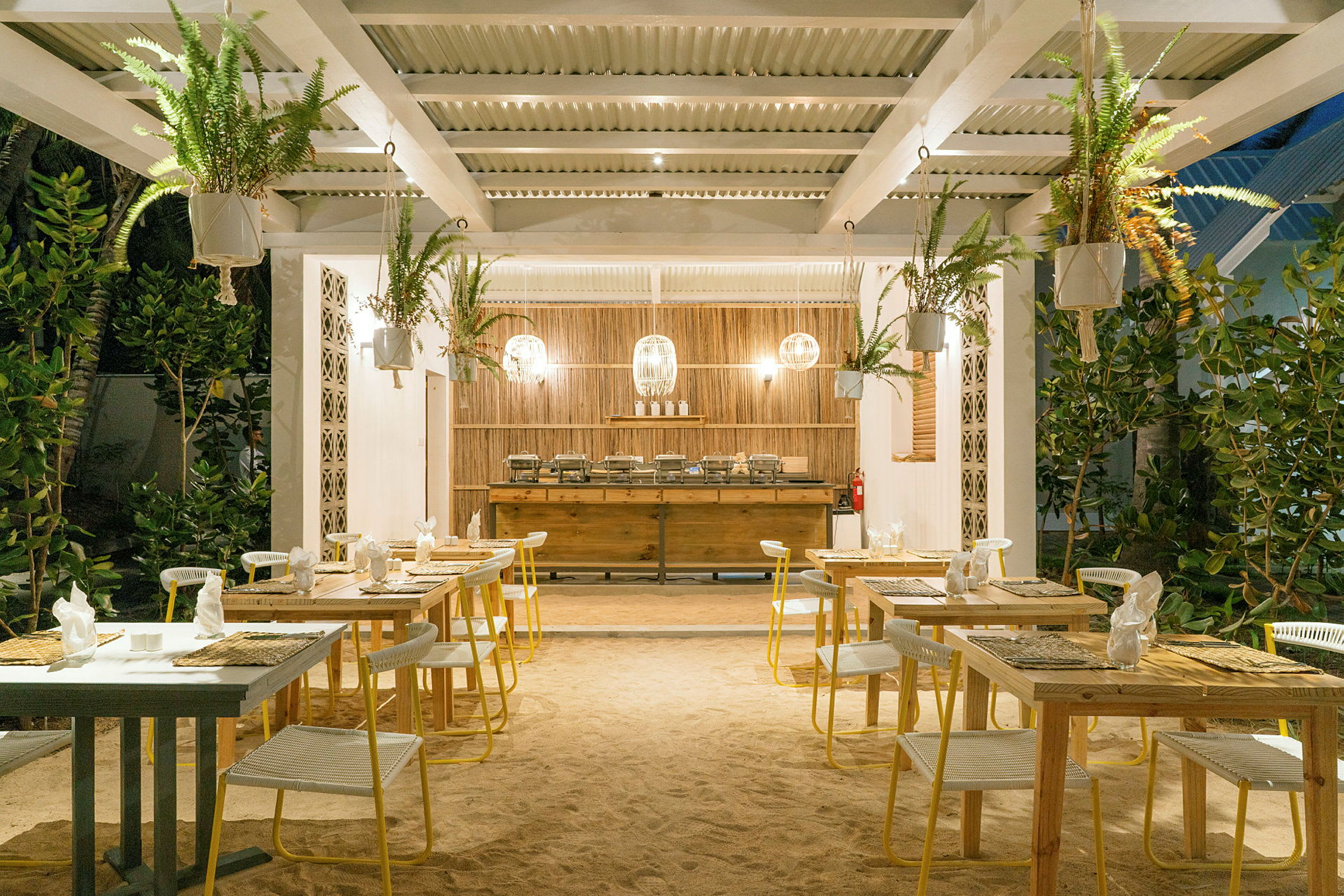 Udendørs restaurant med borde og stole på sandgulv, omgivet af grønt. (AI-genereret tekst)