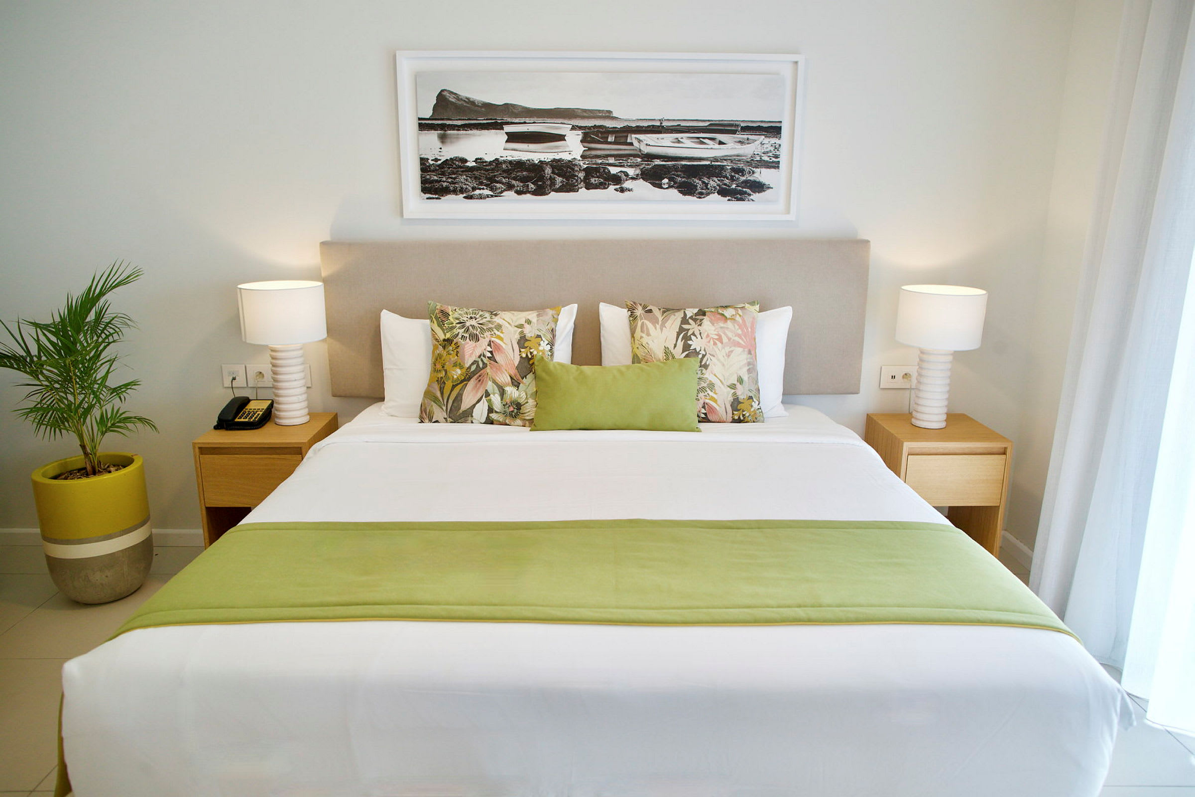 Et hotelværelse med en stor dobbeltseng dekoreret med puder i forskellige farver og mønstre. På hver side af sengen er der et natbord med lampe, og der hænger et billede over sengegavlen. En grøn plante i potte står ved siden af sengen. (AI-genereret tekst)