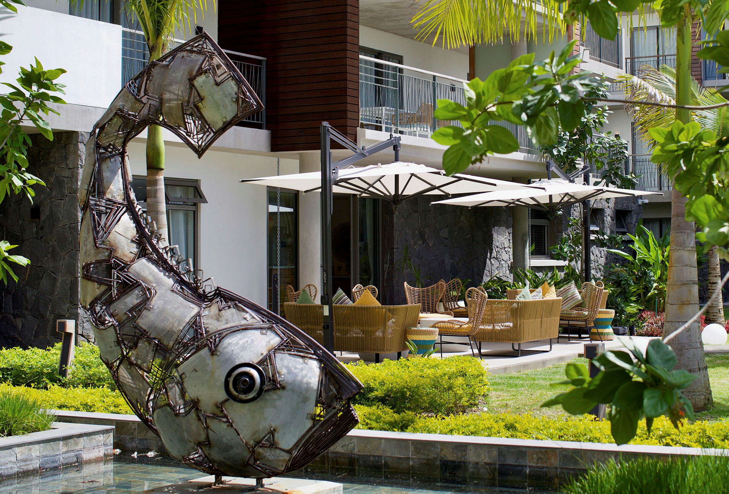 Metalskulptur formet som en fisk foran en terrasse med parasoller og siddegrupper. (AI-genereret tekst)